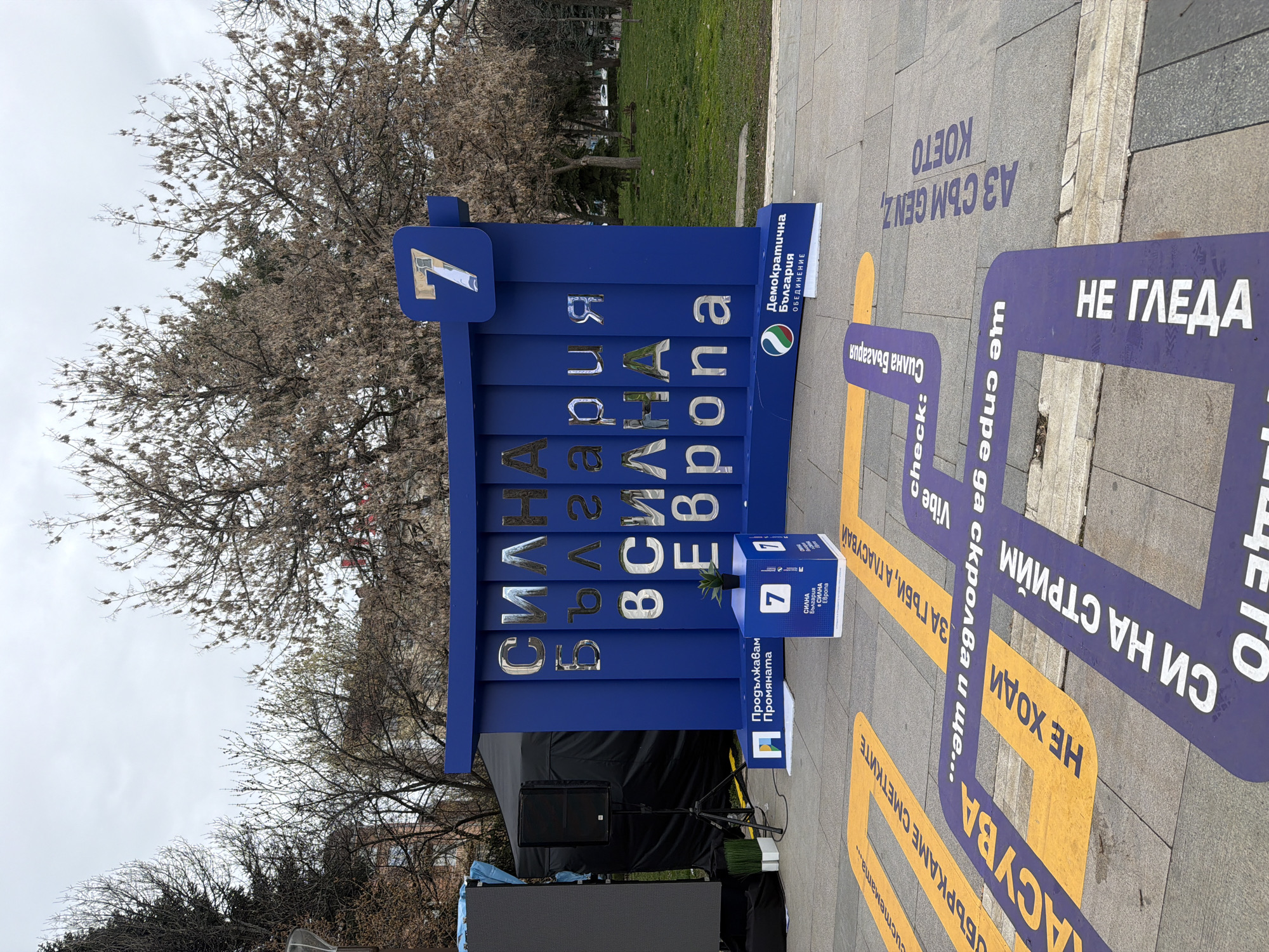 The image depicts an outdoor political event in Sofia, Bulgaria, likely a campaign rally or public relations event for a political party. A large, blue sign reading "Силна България в Силна Европа" (Strong Bulgaria in a Strong Europe) stands prominently. Above the sign is the number seven. The logo for "Демократична България Обединение" (Democratic Bulgaria Union) is also present, indicating the political party involved.
On the ground are painted messages in Bulgarian, appearing like a path. The messages, which include phrases like "не гледа" (doesn't look), "Аз съм Gen Z, което" (I am Gen Z, who), and "За гъби, а гласувай" (Go for mushrooms, but vote), are likely aimed at engaging younger voters.
A black stage-like setup with speakers is behind the sign, suggesting a formal event setup. A small blue box with the number seven on it sits on a small table in front of the large sign, and a small plant sits on top of the small box.
The environment includes a park or public space with bare trees in the background, indicating it's likely early spring or late fall. The image is taken at ground level.