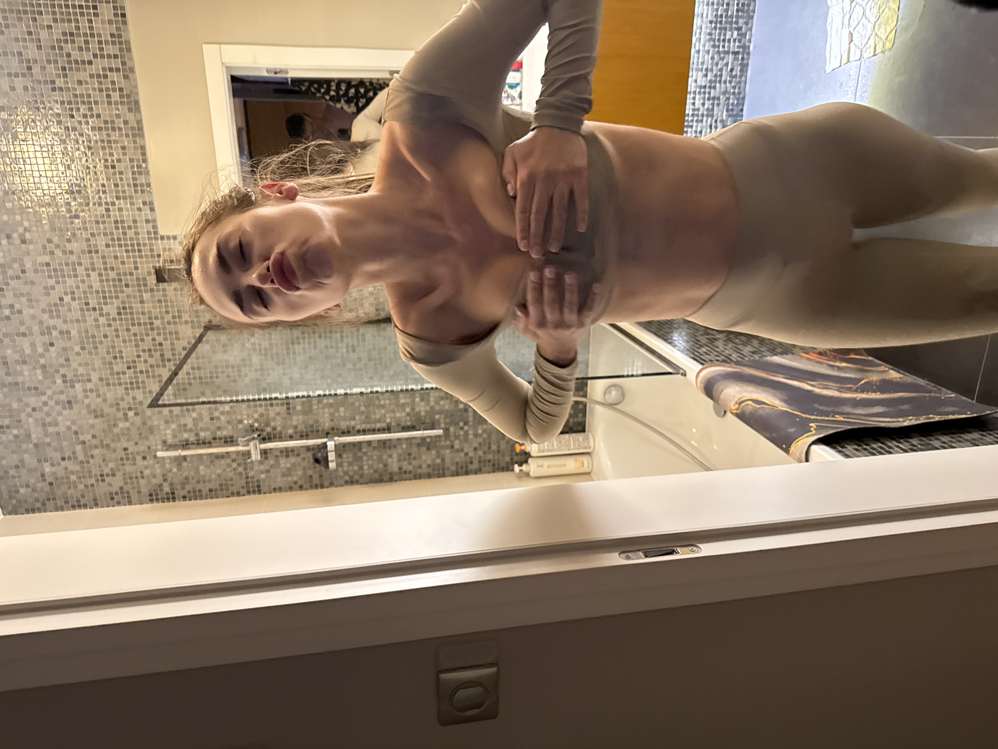The image shows a person in Warsaw, Poland, in what appears to be a bathroom setting. The individual is wearing a body-fitting garment in a nude or skin-tone color. The person is standing with their hands covering their chest. The background includes mosaic tiles on the wall, a shower fixture, a bathtub, and various bathroom accessories. The lighting is bright.