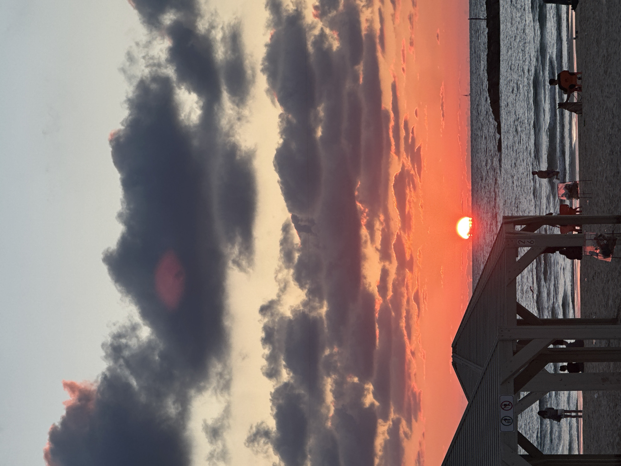 The image shows a sunset at a beach in Tel-Aviv, Israel. In the foreground is a wooden structure, possibly a lifeguard station. The sun is setting over the Mediterranean Sea, with vibrant orange and pink hues illuminating the sky and clouds. Several people are on the beach, either walking, standing, or seated. The sea has moderate waves. Dark, stormy clouds contrast with the bright colors of the sunset in the sky.