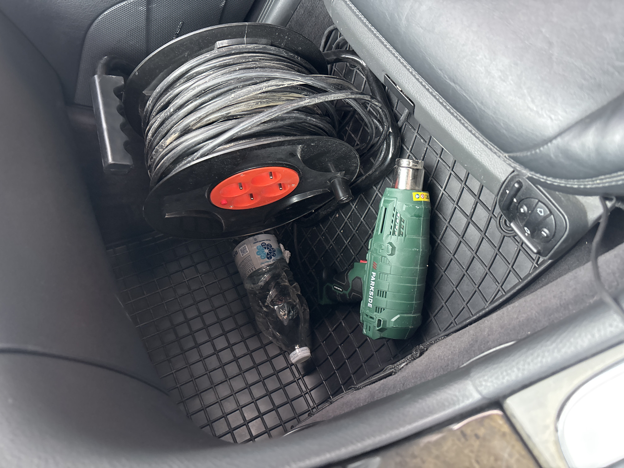 The image shows the interior of a vehicle, likely the footwell of a passenger seat. A black rubber floor mat covers the floor. On the mat, there is a large black cord reel with a red section containing three electrical outlets. A green heat gun, labeled "PARKSIDE," is positioned in front of the cord reel. To the left of the cord reel and heat gun is a clear plastic bottle, possibly containing liquid, with a white cap. The interior of the car appears to be dark-colored, with leather seats visible. The background suggests the car is stationary. There are no people present in the image. The scene is forensic in nature, providing a detailed snapshot of objects within the vehicle. The location provided, Yoakim Gruevo, Bulgaria, is noted as the context for this scene.