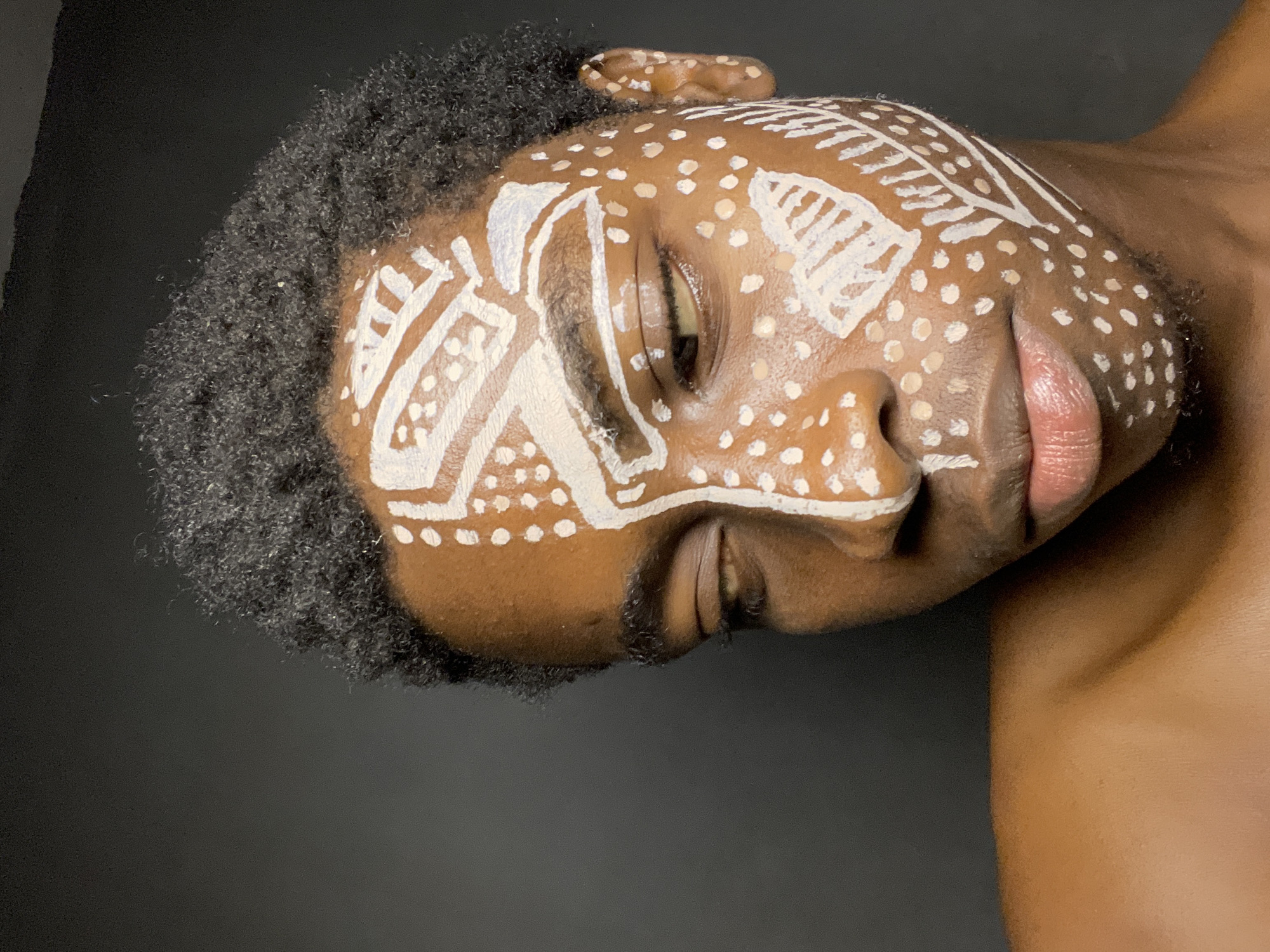 The image shows a close-up of a person's face with white face paint. The paint consists of tribal-like patterns and dots. The person has dark skin, dark curly hair, and is positioned against a dark background. The image does not provide any specific information to identify the location in Nkpor, Nigeria.