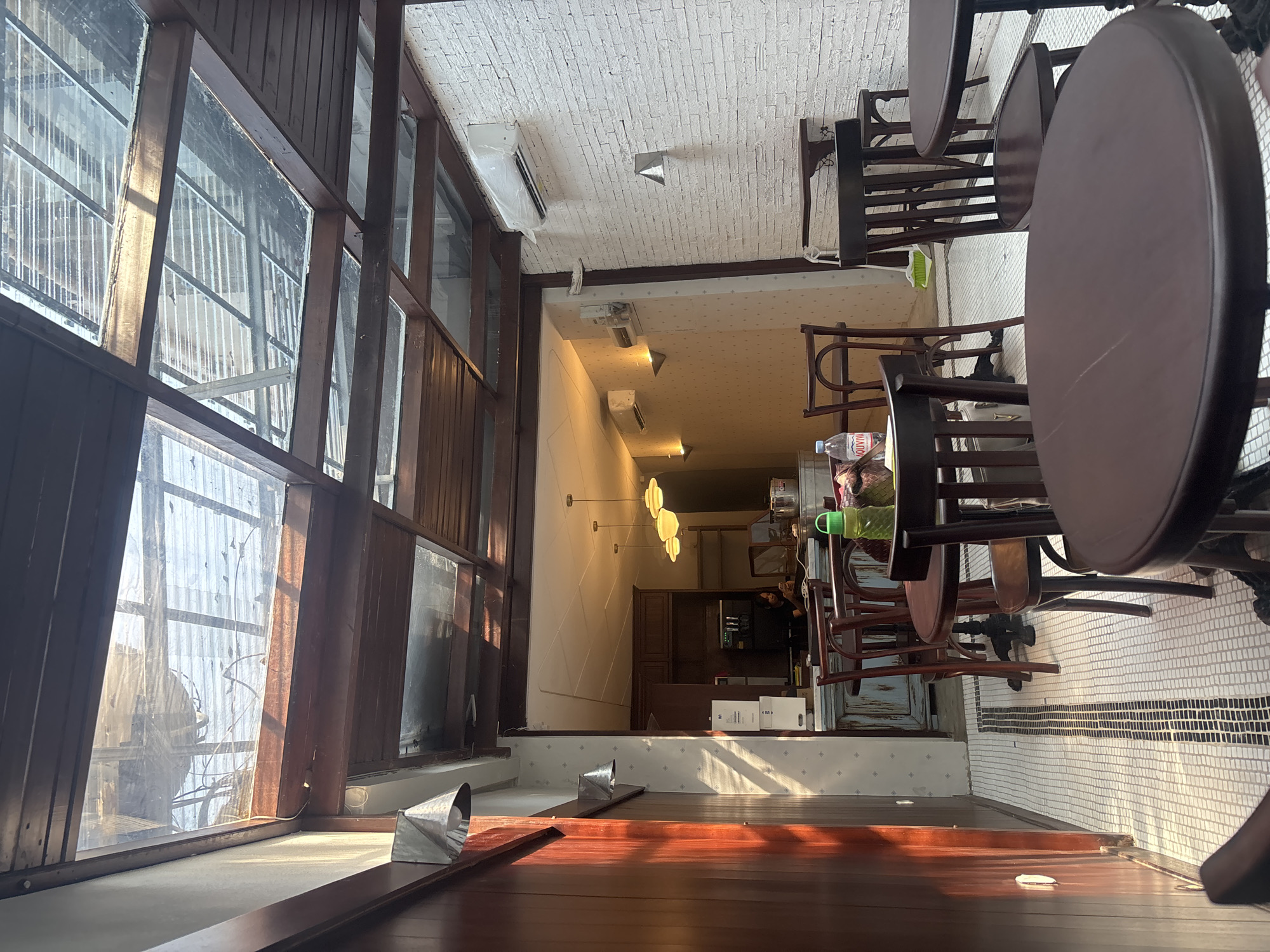 This is an interior shot of a cafe, likely located in South Jakarta, Indonesia, judging by the context. The scene features a courtyard-like area with a transparent corrugated roof that allows natural light to filter through, creating a bright and airy atmosphere. Several dark wooden tables and chairs are arranged throughout the space, some with a vintage aesthetic. The floor is tiled with a white mosaic pattern and a central dark strip.
In the background, a counter area is visible, possibly part of the cafe's service area. A woman is seen behind the counter, appearing to be engaged with her phone. The walls of the cafe are a mix of textured white brick on the right and a lighter patterned wallpaper on the left. Lighting fixtures, including modern ceiling lights and wall sconces, are visible, suggesting the cafe is equipped for evening operation. An air conditioning unit is mounted on the brick wall.
The overall impression is of a relaxed and casual cafe setting, perhaps a popular spot for people to gather.
