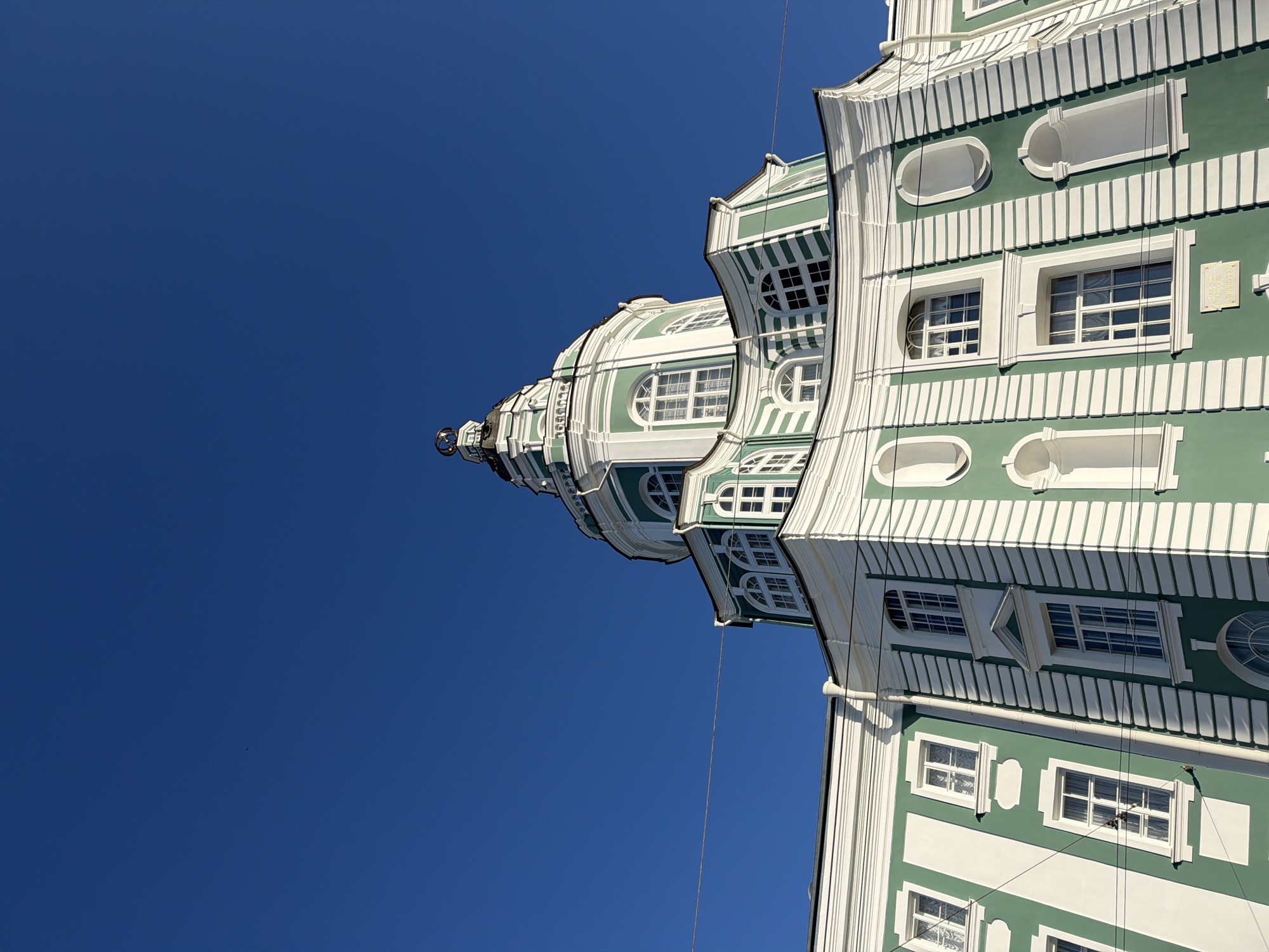 The image shows a low-angle shot of the bright green and white dome and facade of the Smolny Cathedral in Saint Petersburg, Russia, against a clear, vibrant blue sky. The ornate architectural details of the cathedral, including arched windows, decorative moldings, and a prominent spire topped with a golden orb, are clearly visible. The building appears to be in good condition, bathed in sunlight, casting subtle shadows on its exterior. A thin wire runs horizontally across the upper portion of the building and the sky. There are no people or animals visible in the frame. The perspective suggests the photograph was taken from ground level, looking upwards towards the cathedral.