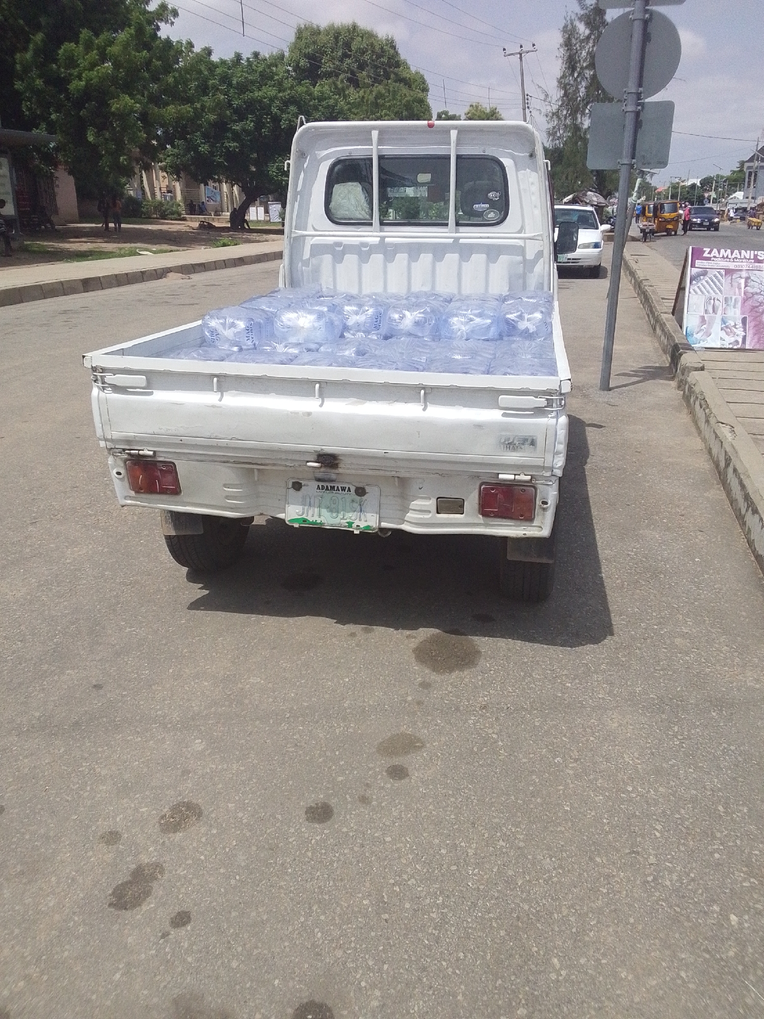 A white pickup truck is parked on the side of a paved road in Jalingo, Nigeria. The truck bed is filled with many clear plastic bottles of water. The license plate on the back of the truck reads "ADAMAWA JHN 919 GK". A street signpost stands to the right of the truck. In the background, there are trees, buildings, and other vehicles, including a white car, a tricycle, and a black car. The sky is overcast.