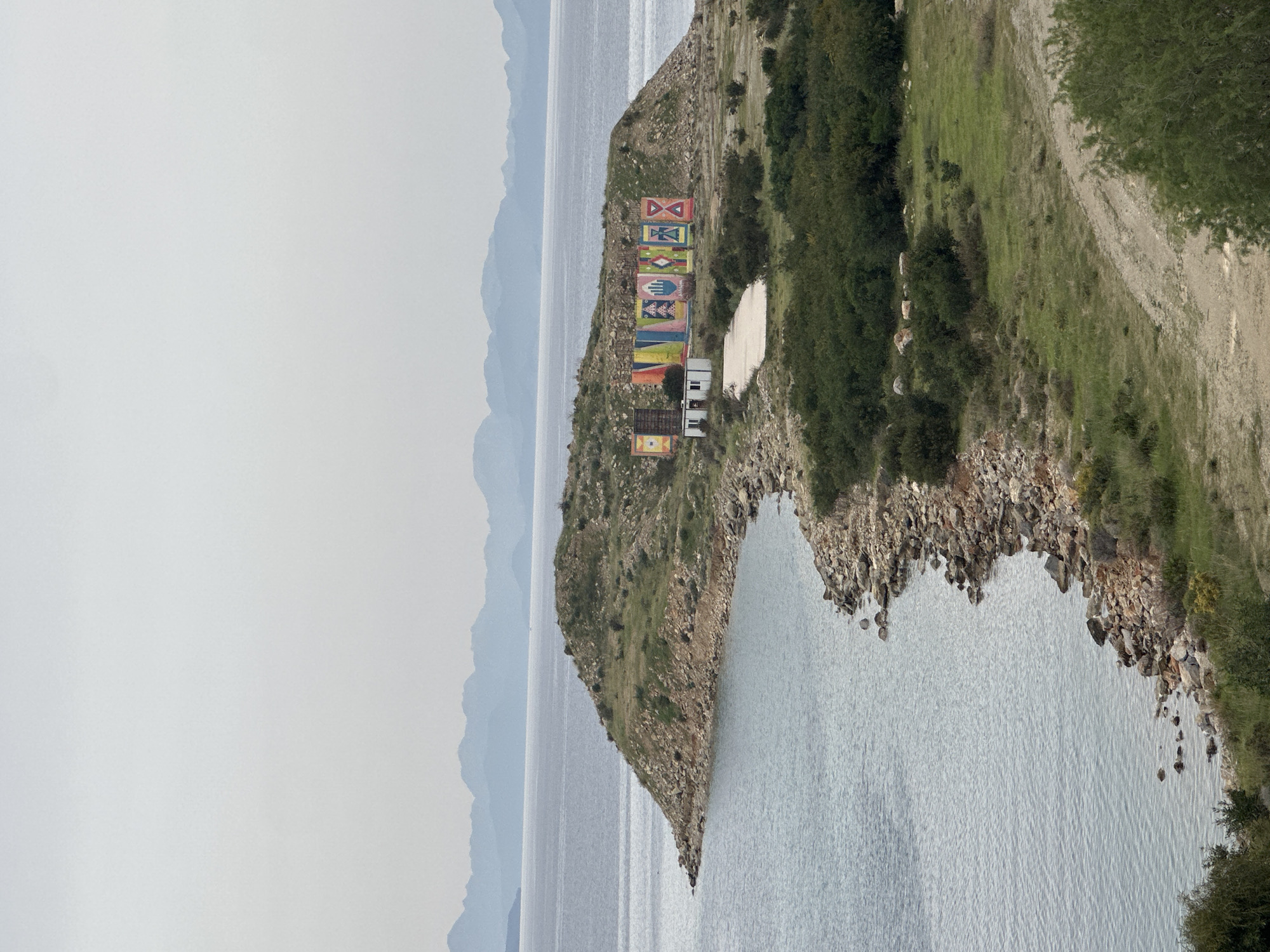 The image shows an aerial, sideways view of a promontory extending into the sea, likely taken in Datça, Turkey. The landscape is dominated by the shoreline, with the clear water stretching into the distance where hazy mountains fade into the sky. The promontory itself is covered in sparse vegetation, dirt, and rocks. At the end of the promontory, closest to the viewer, is a series of eight brightly painted panels displaying various symbols. Behind the panels are a few small white buildings. A dirt path runs along the top of the promontory on the right side of the image, bordering the vegetation. The sky is overcast, contributing to the muted color palette.
