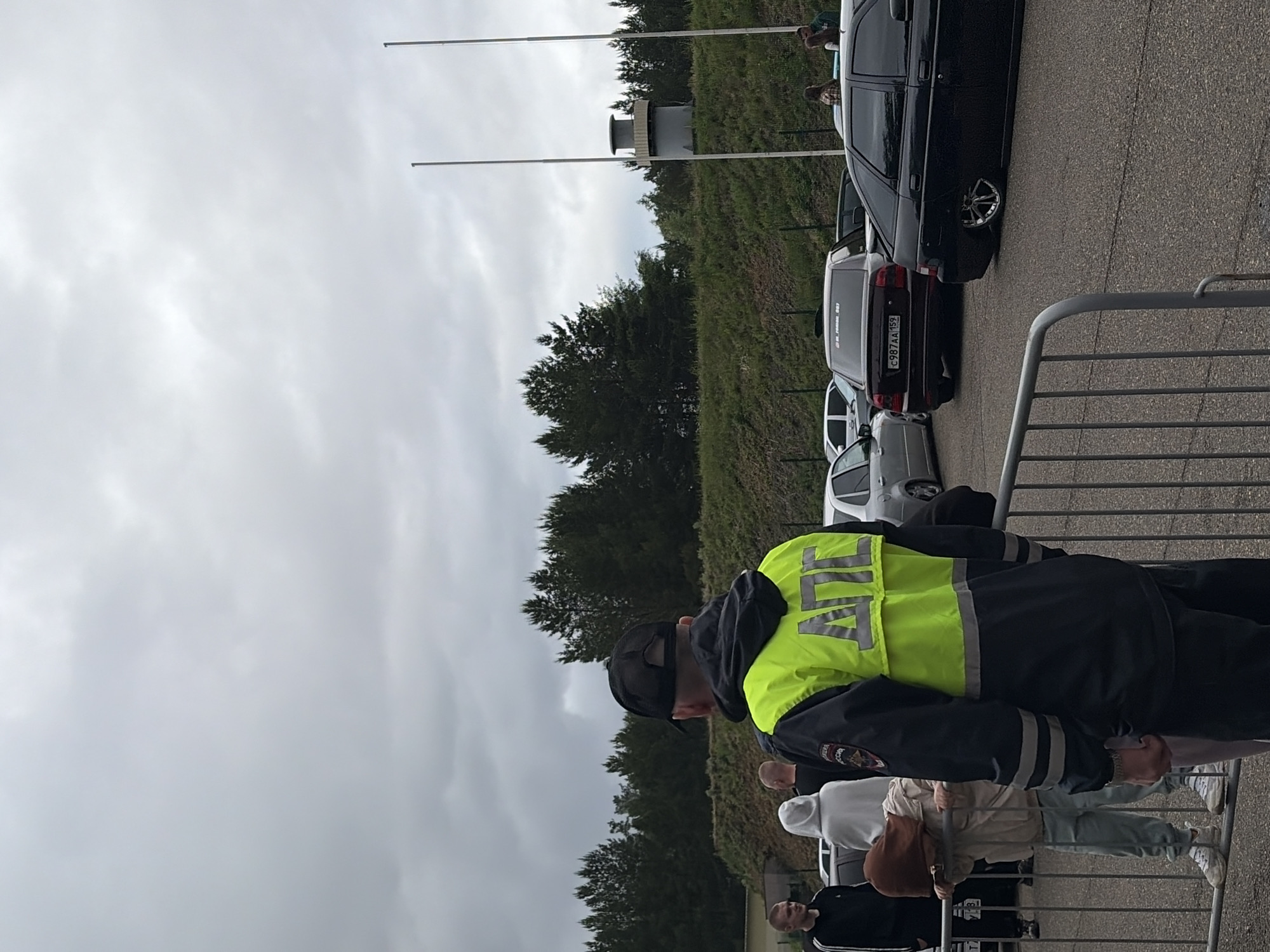 An outdoor scene in Прикамский, Russia, depicts an overcast day with multiple individuals and vehicles on an asphalt surface, bordered by a grassy embankment and trees. In the foreground, a metal barrier extends horizontally. A male figure stands with his back to the camera, wearing a black baseball cap, a black hooded garment, and a reflective neon yellow and dark blue uniform jacket. The jacket features "ДПС" in reflective lettering on the back and a patch with the Russian Federation emblem on the left sleeve. To his left, another male in a black shirt with white sleeve stripes is visible, along with a person in a white hoodie and a third person in a light brown hooded jacket and light blue pants, leaning over the metal barrier.
Multiple vehicles are present on the asphalt. Prominently featured is a dark red sedan, identified as a Lada Priora or similar model, with the Russian license plate C987AA 159. The rear window displays text, partially visible as "M_ROMAN_003". To its right, a black sedan, also a Lada model, exhibits a significantly lowered suspension and custom silver multi-spoke wheels. Other less distinct silver and dark vehicles are visible further back.
The background consists of a sloping green and brown grassy embankment topped with a dense line of coniferous trees. Above the tree line, two tall, slender, light-colored poles stand vertically. Between these poles, on top of the embankment, a grey, cylindrical concrete structure with a slightly wider, flat top is present, resembling an observation tower or a specific industrial structure. The sky is uniformly grey with dense cloud cover.