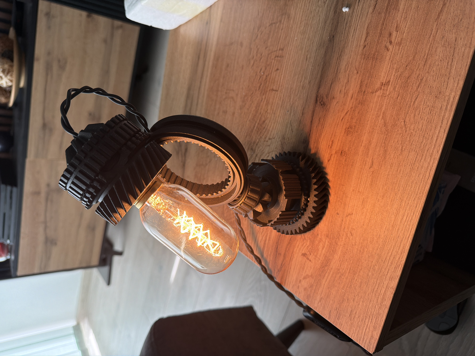 The image, presented in a vertical orientation, captures a distinctive desk lamp casting a warm glow on a wooden surface. The lamp exhibits an industrial or steampunk design, constructed from dark-colored components that resemble intermeshing gears and mechanical parts, possibly indicating a 3D-printed creation. An illuminated Edison-style filament bulb, with its intricate coiled filament, emits a bright, yellow-orange light, serving as the central light source. A black, twisted electrical cord extends from the lamp, leading out of the frame towards the bottom right.
The wooden surface beneath the lamp appears to be a desk or table, showcasing a pronounced wood grain pattern. The warm light from the bulb intensely illuminates the area to the right of the lamp, creating a strong contrast. In the upper left corner of the frame, glimpses of another wooden surface and dark shelving are visible, alongside a blurred white object. The bottom left section of the image reveals a light-colored floor and a hint of a wall or window.
No specific activity is taking place other than the lamp being lit, suggesting its functional use as a decorative light source. The warm illumination contributes to a cozy and inviting atmosphere. The time of day is not explicitly clear from the image, but the reliance on the lamp for light implies it could be evening or an indoor setting with limited natural light. There is no visible text within the frame, and no specific visual cues to indicate the stated location of Sofia, Bulgaria.