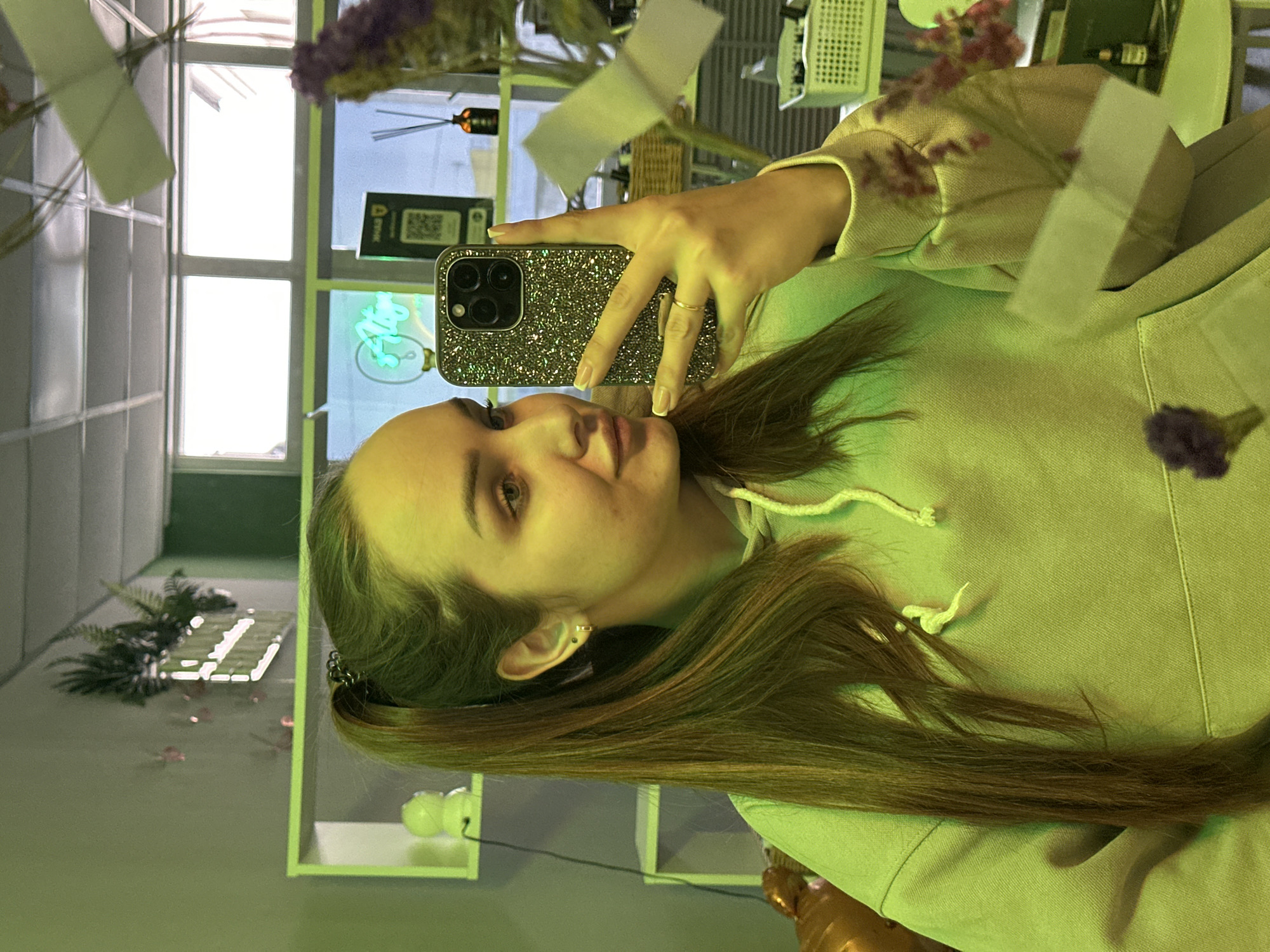 A woman is taking a selfie with an iPhone in a green-lit room. She has light brown hair and is wearing a light green hoodie. Her phone has a glittery case. The location is Chaikovsky, Russia, as identified by a QR code visible on a sign in the background. The room contains decorative plants, shelves with objects, and a window with a view of the outside.