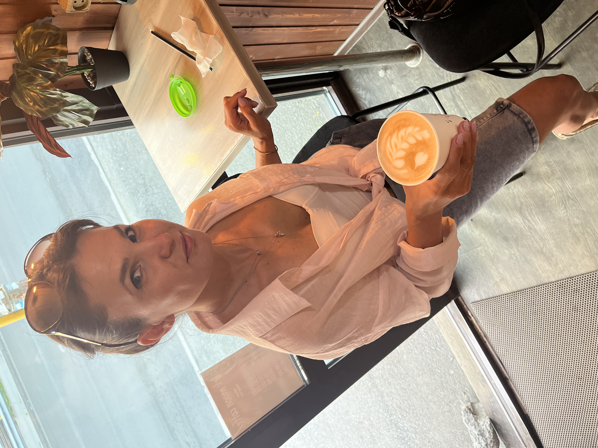 A woman is seated at a table in a cafe, holding a coffee cup with latte art. She is wearing a light pink button-down shirt over a white top and denim shorts. Sunglasses are perched on her head. The cafe appears to be located near a window overlooking a street, suggesting it is in a city. The environment includes a wooden table with a green condiment holder, a straw, a napkin, and a potted plant. The floor is a light wood laminate, and part of a chair is visible. The cafe's interior design features wooden paneling. The presence of a sign visible through the window, which appears to be in Cyrillic, suggests the location is in Russia. Specifically, the context indicates this scene is in Petropavlovsk-Kamchatsky, Russia.