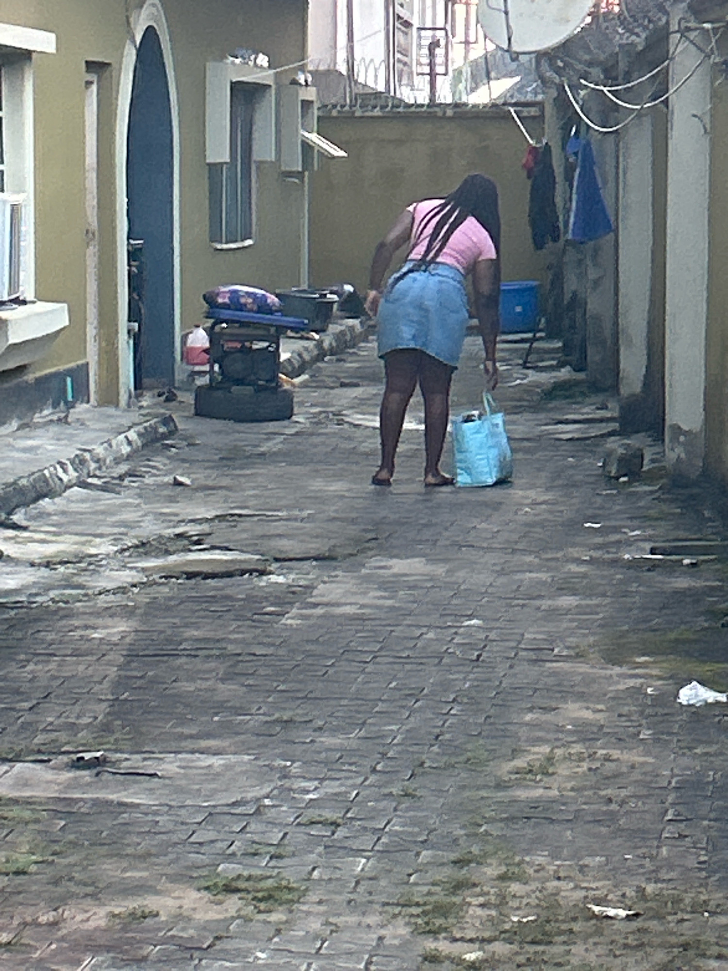 In Ikotun, Nigeria, a woman with long black hair, wearing a pink t-shirt and a denim skirt, bends over to pick up a light blue shopping bag from a cobblestone pathway. The pathway is cracked and uneven, with some patches of green vegetation growing through. To the left of the pathway, a light yellow building features a dark blue arched doorway and a white-framed window. Further down the pathway, a generator is visible, topped with a blue and white patterned mat. The background shows more buildings and overhead wires.