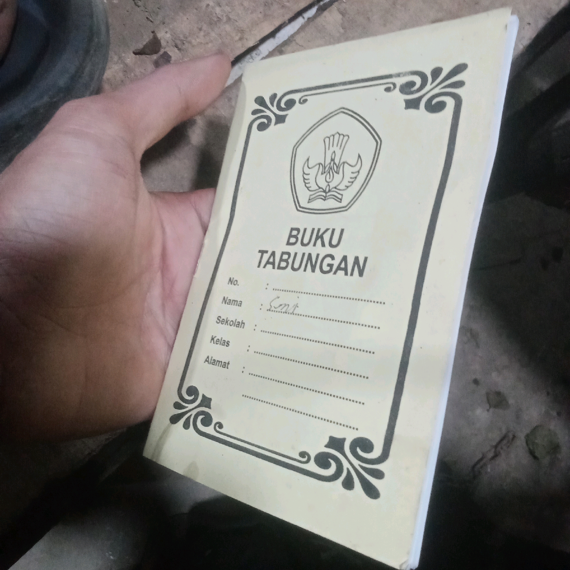 A hand is holding a light yellow booklet with a stylized emblem and decorative borders. The booklet has text printed in black, with lines for filling in information. The printed text reads "BUKU TABUNGAN" (Savings Book) at the top. Below this, there are fields for "No.", "Nama" (Name), "Sekolah" (School), "Kelas" (Class), and "Alamat" (Address), each followed by a dotted line for writing. The "Nama" field has handwritten text that appears to be "Sut". The booklet is positioned at an angle, partially obscuring the background, which appears to be a textured, dirty surface, possibly concrete or stone, with some debris scattered around. The lighting suggests an indoor or dimly lit outdoor environment. There are no discernible landmarks that would indicate a specific location in Karawang, Indonesia. The image is a close-up shot focused on the booklet and the hand holding it.