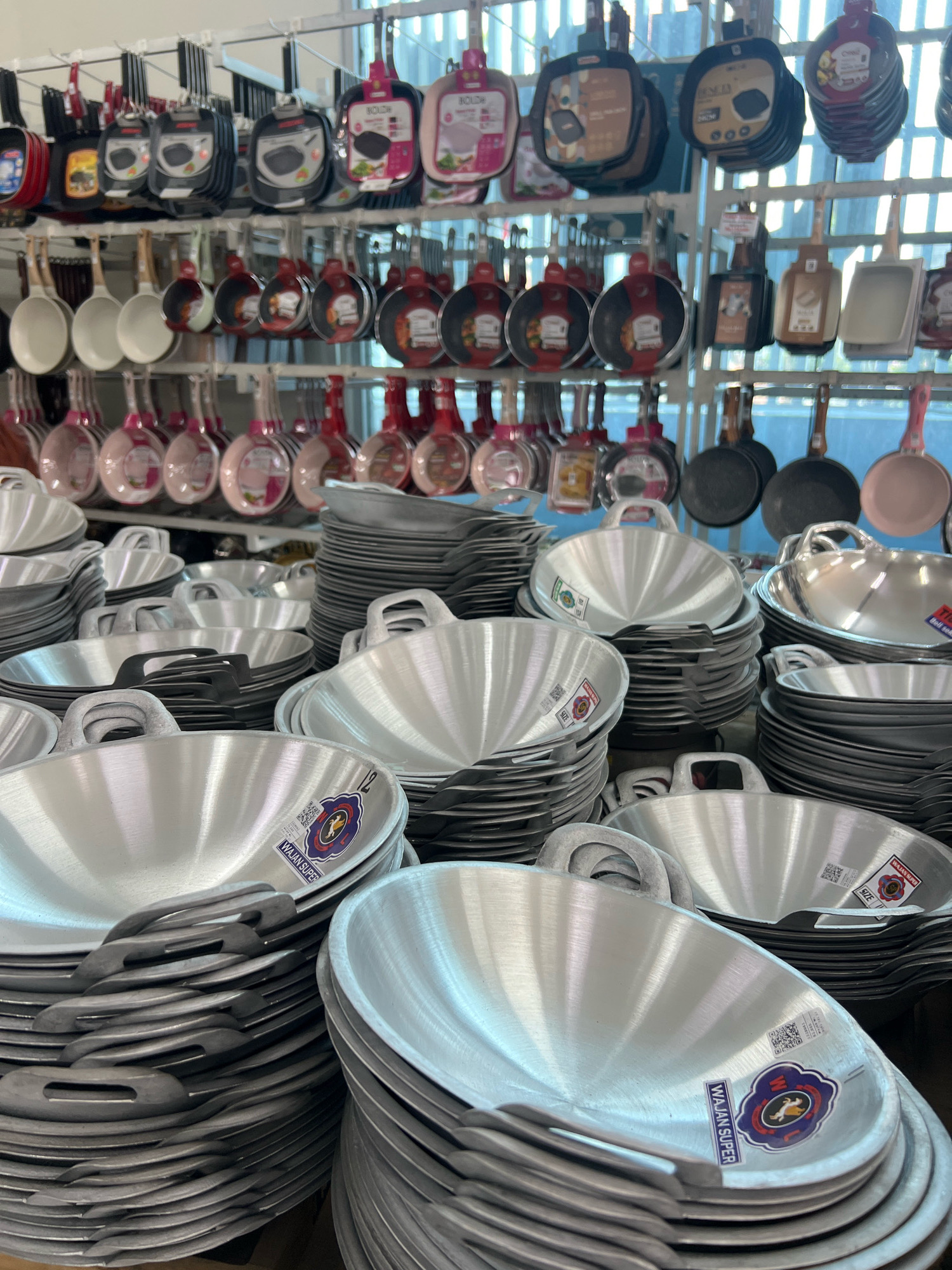This is a detailed, forensic, matter-of-fact description of a store display of cookware in Yogyakarta, Indonesia. The scene is an interior retail environment, likely a kitchenware store, with rows and stacks of various types of pots and pans. **Subjects and Objects:** * **Cookware:** The primary objects are numerous pots and pans, predominantly made of aluminum and possibly some non-stick materials. They are displayed in various ways: * **Hanging Displays:** On overhead racks, several rows of pans are hung by their handles. These include flat-bottomed frying pans, possibly woks, and smaller saucepans. Some are in packaging, with colorful labels and images. Examples of visible labels include "HOLIDA" and others with graphics of food. * **Stacked Displays:** Large quantities of metal woks or deep frying pans are meticulously stacked in neat columns on shelves or display units. These stacks are dense, with the individual pans partially visible. The handles are a prominent feature, appearing to be made of metal. * **Individual Pans:** Some individual pans are also visible, showcasing their shapes and finishes. Many of the stacked aluminum pans have labels attached to their interiors, with text such as "WAJAN SUPER," "SIZE M," and QR codes. The labels feature a blue and red emblem with a horse or animal graphic. * **Color Palette:** The dominant colors are metallic silver (from the aluminum cookware), black (from some pans and handles), and various colors from the packaging and labels (pink, red, orange, blue, white, and brown). **Environment:** * **Retail Setting:** The presence of organized displays, overhead racks, and shelves indicates a commercial retail space. * **Lighting:** The scene appears to be well-lit, with natural light possibly filtering in from a background, suggested by the bright, diffused light in the upper sections of the image. * **Background:** The background shows a wall or structure with vertical elements, possibly shelving supports or the building's framework, behind the cookware displays. The upper part of the image reveals a light-colored, possibly translucent material, suggesting it could be a window, skylight, or part of the store's ceiling structure. **Actions and Interactions:** * There are no people visible in the image, so no actions or interactions between individuals are present. The scene is solely focused on the display of merchandise. **Location Context (Yogyakarta, Indonesia):** * The presence of the brand names and product types (e.g., "Wajan" which is the Indonesian word for wok or frying pan) strongly suggest a retail