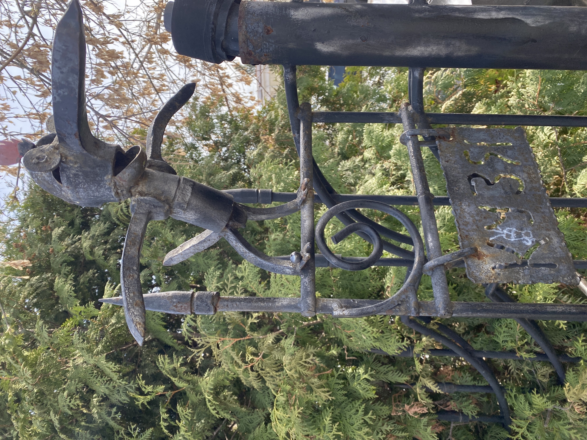 This image shows a fragment of a metal fence in Yekaterinburg, Russia. The fence features decorative elements including spear-shaped spikes and a curly design. A rusty metal plate is attached to the fence, bearing illegible cut-out letters or numbers. The fence is set against a backdrop of green coniferous trees. The overall scene suggests an outdoor setting with aged or weathered metalwork.