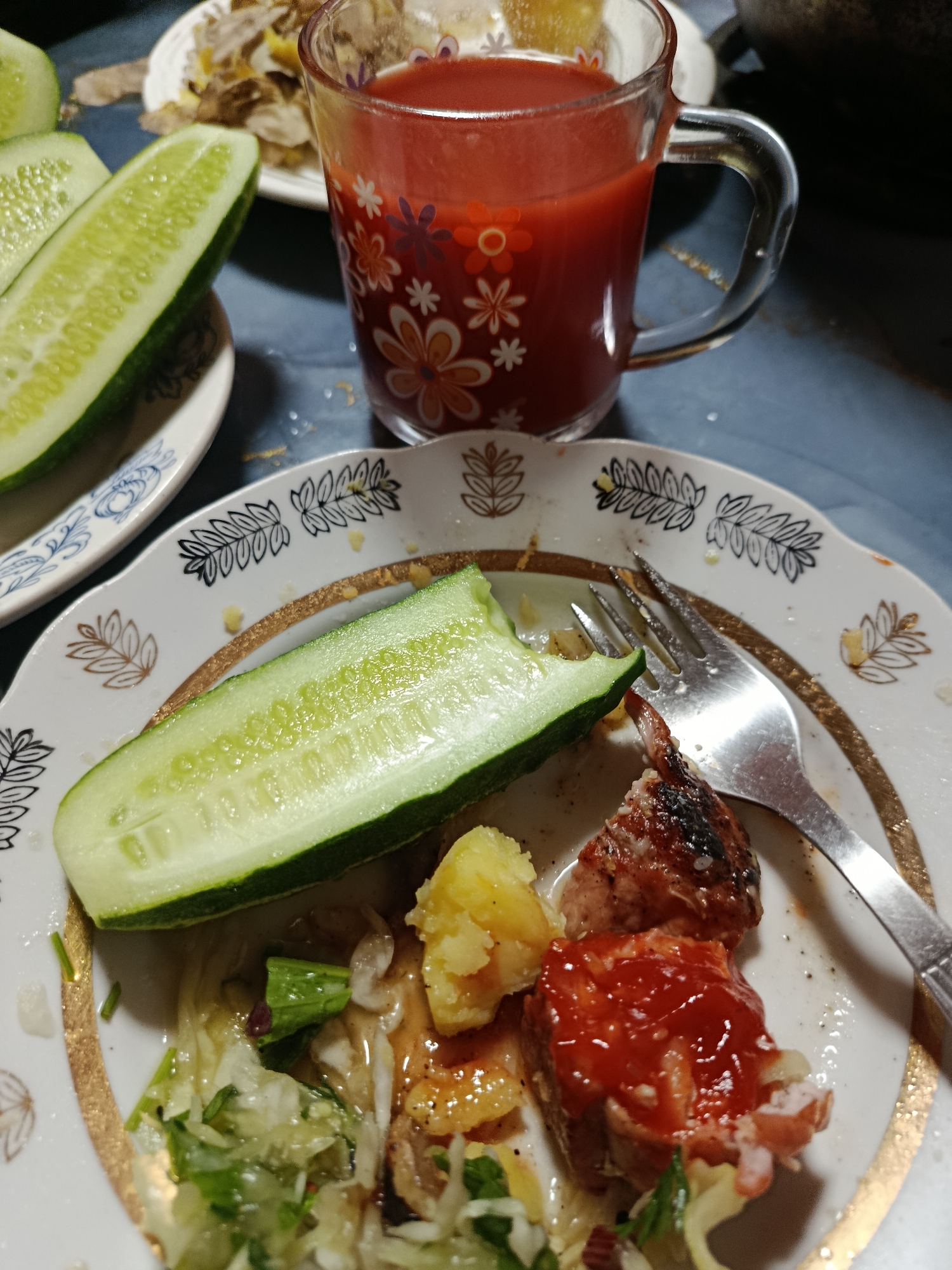 In an indoor setting, a plate of food sits on a dark blue tablecloth. The plate, decorated with a gold rim and black floral patterns, contains a half-eaten meal. Visible on the plate are pieces of meat covered in red sauce, sliced potatoes, shredded salad, and a large slice of cucumber. A fork rests on the plate, next to the food. To the left of the plate, another slice of cucumber is positioned on a small, white dish with a blue floral design. In the background, a glass mug filled with a red beverage, possibly juice, is adorned with a floral pattern of white and orange flowers. The overall scene depicts a meal that has been partially consumed, with the food arranged in a somewhat messy fashion.