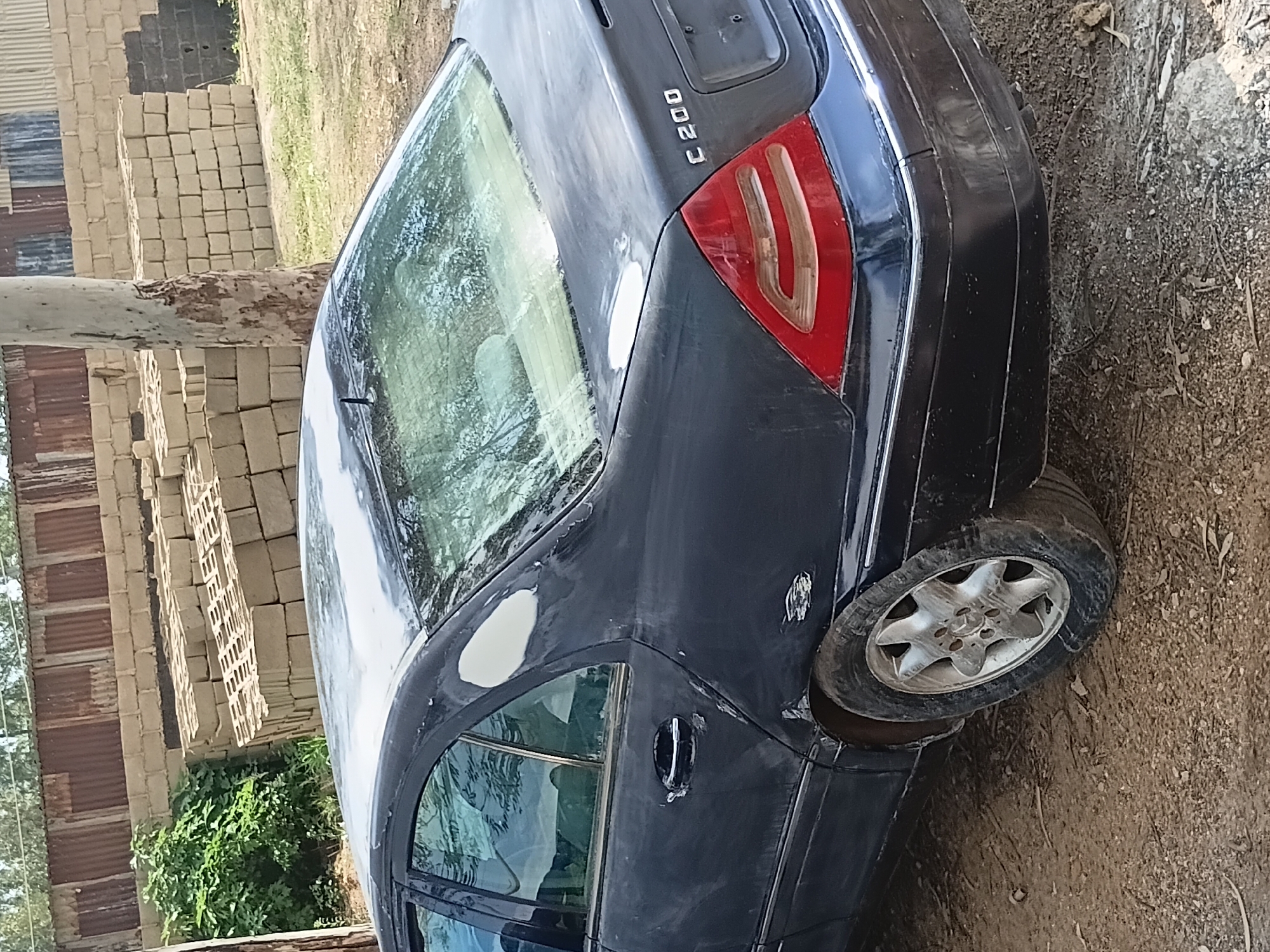 The image shows the rear passenger side of a dark-colored sedan, likely a Mercedes-Benz C200, parked outdoors on a dirt and gravel surface. The car appears to be in a state of disrepair or renovation. Patches of primer or body filler are visible on the rear fender and trunk lid, indicating bodywork is in progress. The paint is dull and appears scuffed in places. The rear taillight is visible, as is the C200 badging. The car's wheel is a silver multi-spoke design. In the background, there is a stack of gray cinder blocks and a rustic fence made of corrugated metal and wooden posts. Lush green foliage is visible to the left of the car, and a large tree trunk is to the right, partially obscuring the background. The overall scene suggests a utilitarian or storage area.