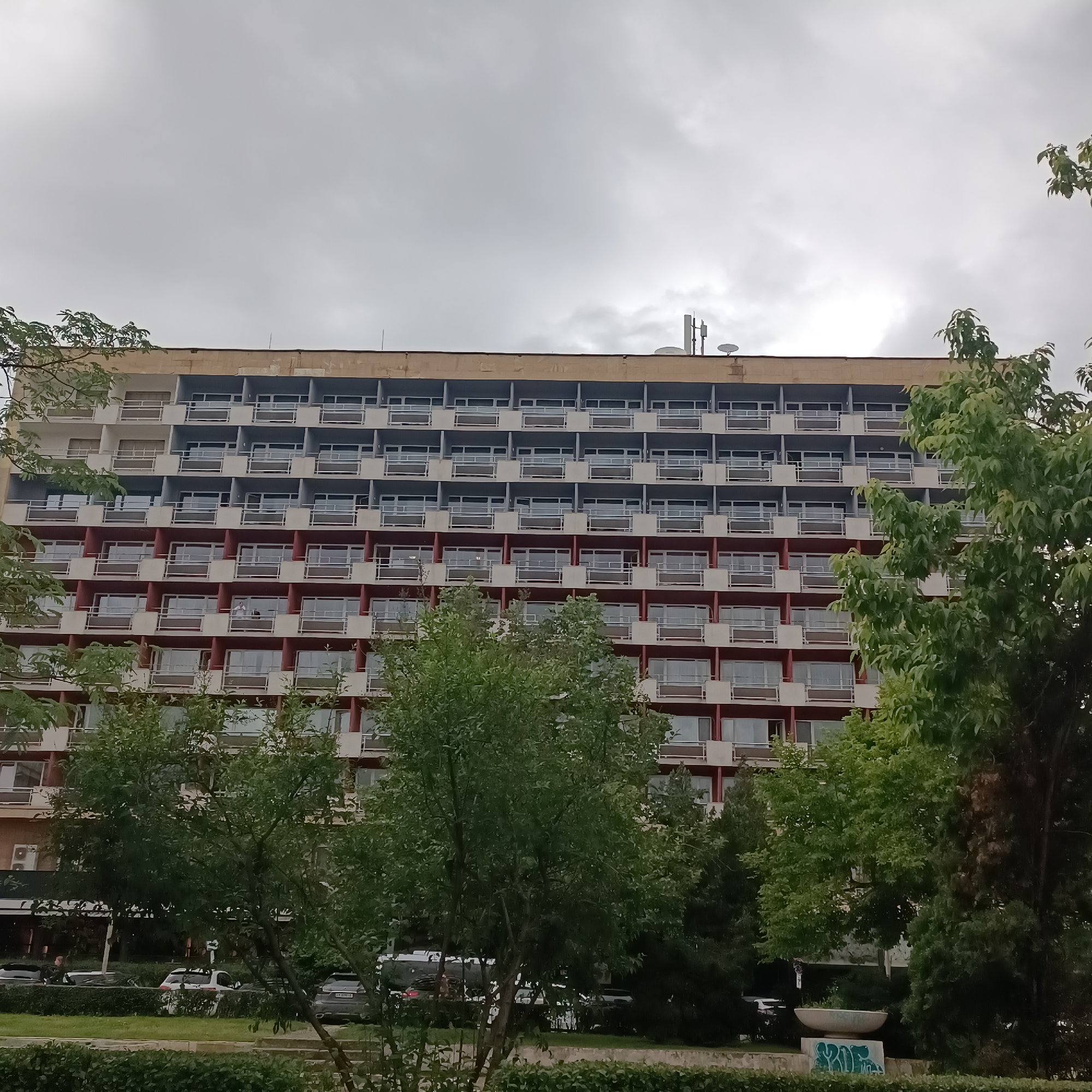 This image captures an urban scene in Sofia, Bulgaria, dominated by a large, multi-story building under an overcast sky. The building, likely a hotel or residential complex, features a repetitive pattern of balconies across its facade. The upper floors show sections with light gray or blue walls, while the lower visible floors feature red or brown wall sections between the balconies. Each balcony has a railing, and the windows behind them reflect the cloudy sky. Antennas and satellite dishes are visible on the building's rooftop.
Dense green trees and bushes partially obscure the lower and side portions of the building, creating a layered foreground. One person can be faintly seen standing on a balcony on one of the mid-level floors. At the very bottom, partially hidden by the foliage, a street with several parked or moving cars is visible.
The weather appears to be cloudy, with a uniform gray sky suggesting an overcast day without direct sunlight. On a concrete planter or low wall in the lower right foreground, green graffiti is visible, which appears to spell out letters such as "SKEP" or "SKOP" amidst other markings. The overall scene is typical of a bustling city environment, with architectural elements complemented by urban greenery.