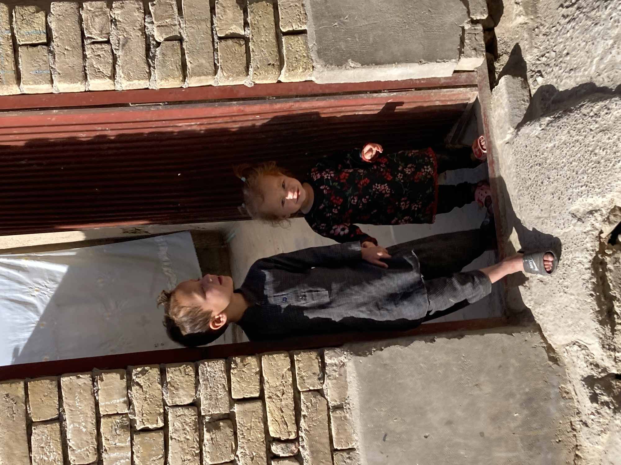 Two children, a boy and a girl, are positioned in what appears to be a recessed area or doorway of a building. The building appears to be made of concrete and brick. The area is partially enclosed by a red frame. The boy is wearing dark clothing and sandals; the girl is wearing a long-sleeved floral patterned dress and sandals. The setting appears to be an urban environment, given the materials of construction. The location is Quetta City Tehsil, Pakistan.