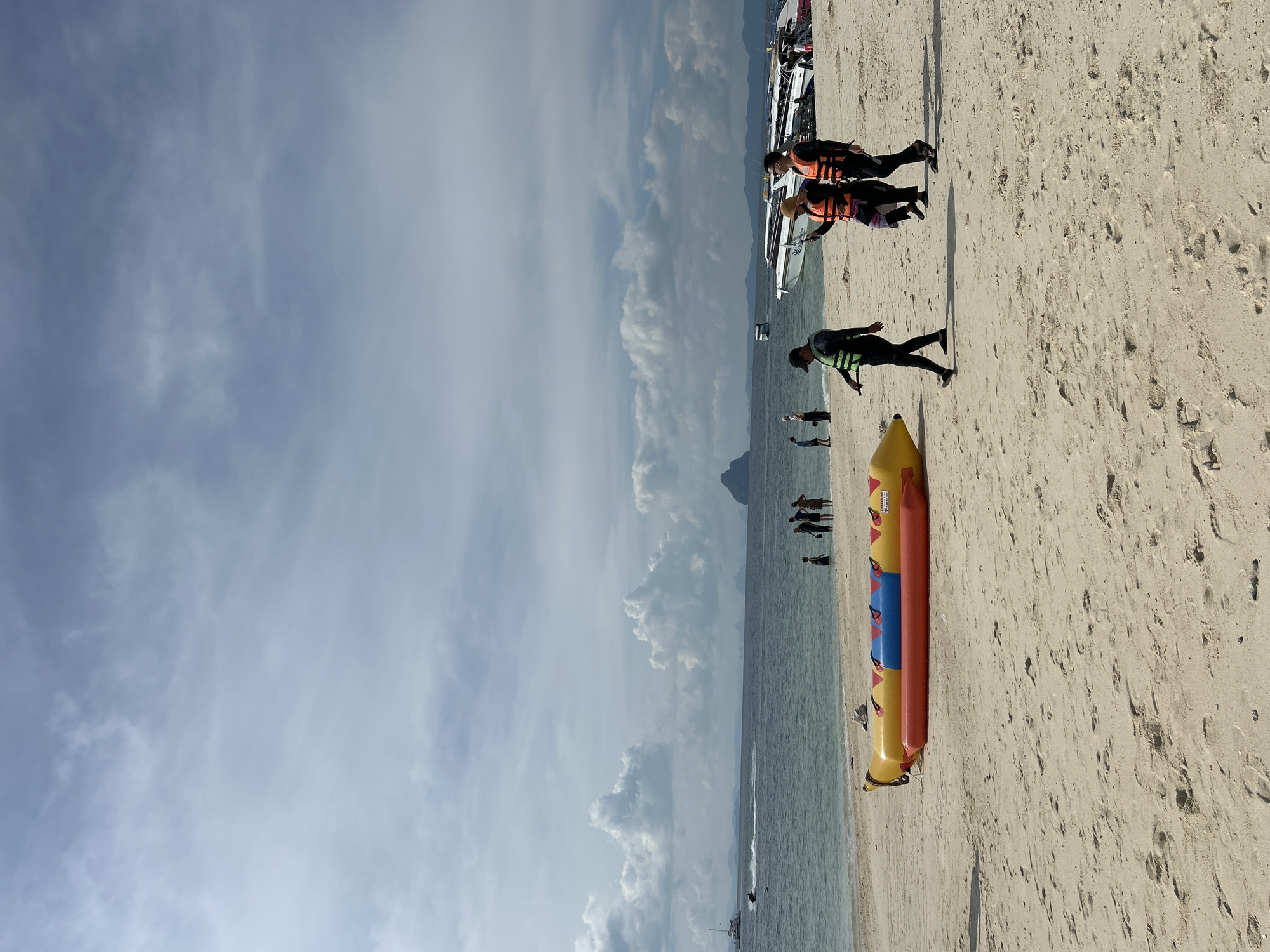 This is a full, eye-level, outdoor shot of a sandy beach in Phru Nai, Thailand, with the ocean and sky in the background. In the foreground, there is a deflated yellow and red inflatable banana boat lying on the sand. Three people in orange life vests are walking from left to right across the beach. One person, wearing a black cap and a black vest, walks in front and to the left of the other two. The other two people, a woman and a man, follow closely behind. In the mid-ground, several other people are standing in the shallow water. The ocean extends to the horizon, where a few small islands are visible. Several boats are docked in the water. The sky is cloudy with patches of blue.