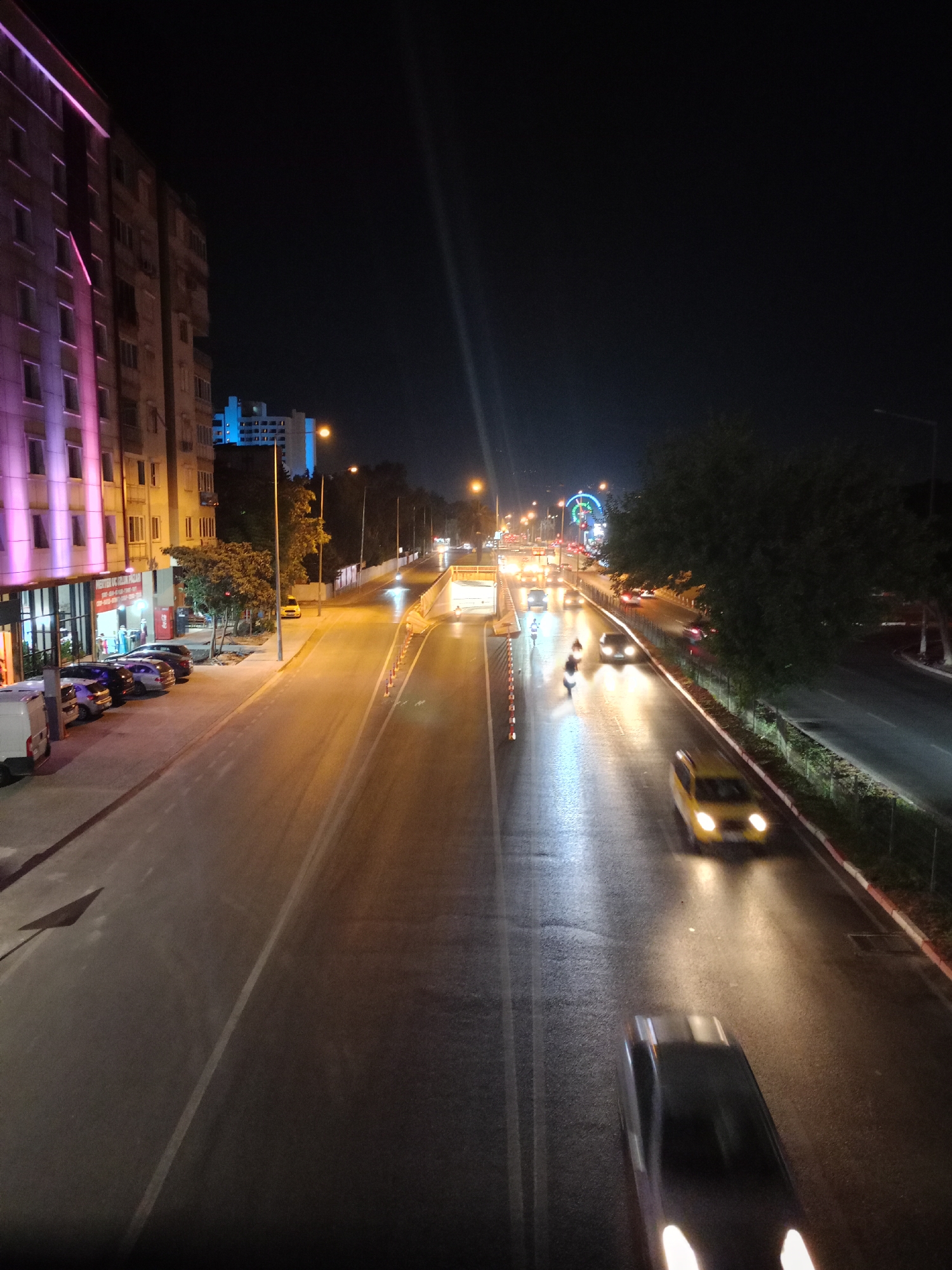 The image captures a nighttime view of a multi-lane road in Antalya, Turkey, likely near the city center or a busy intersection. On the left, a tall, multi-story building is illuminated with purple and pink lights, suggesting it is a commercial or residential structure. In front of this building, several cars are parked along the sidewalk, and a white van is also visible.
The road itself is wet, reflecting the streetlights and the headlights of passing vehicles. Traffic cones are placed along the road, indicating a temporary lane closure or construction. Several cars and motorcycles are in motion on the road. In the distance, a Ferris wheel-like structure is illuminated with colorful lights, hinting at an amusement park or a festive area. Trees are present on the right side of the road, casting shadows and adding to the urban landscape. Streetlights are positioned along the road, providing illumination for the scene. The overall atmosphere is that of a vibrant, active city at night.