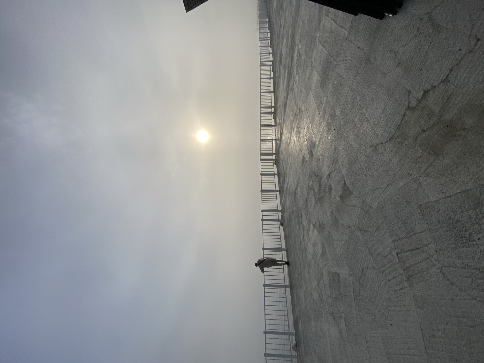 A lone person stands at a white railing overlooking a foggy expanse of water, likely Lake Baikal, near the city of Listvyanka, Russia. The sun is a diffused bright orb in the hazy sky. The foreground is a grey concrete patio with a rough, textured surface, appearing wet in places, reflecting the low light. The railing runs horizontally across the midground, separating the patio from the water. The figure, seen from behind, is wearing light-colored shorts and a light-colored shirt. Their posture suggests contemplation or observation of the water and the fog. To the far right, a dark, angular object, possibly part of a structure, juts into the frame from the edge of the image. The overall atmosphere is serene and slightly melancholic due to the fog and muted light.