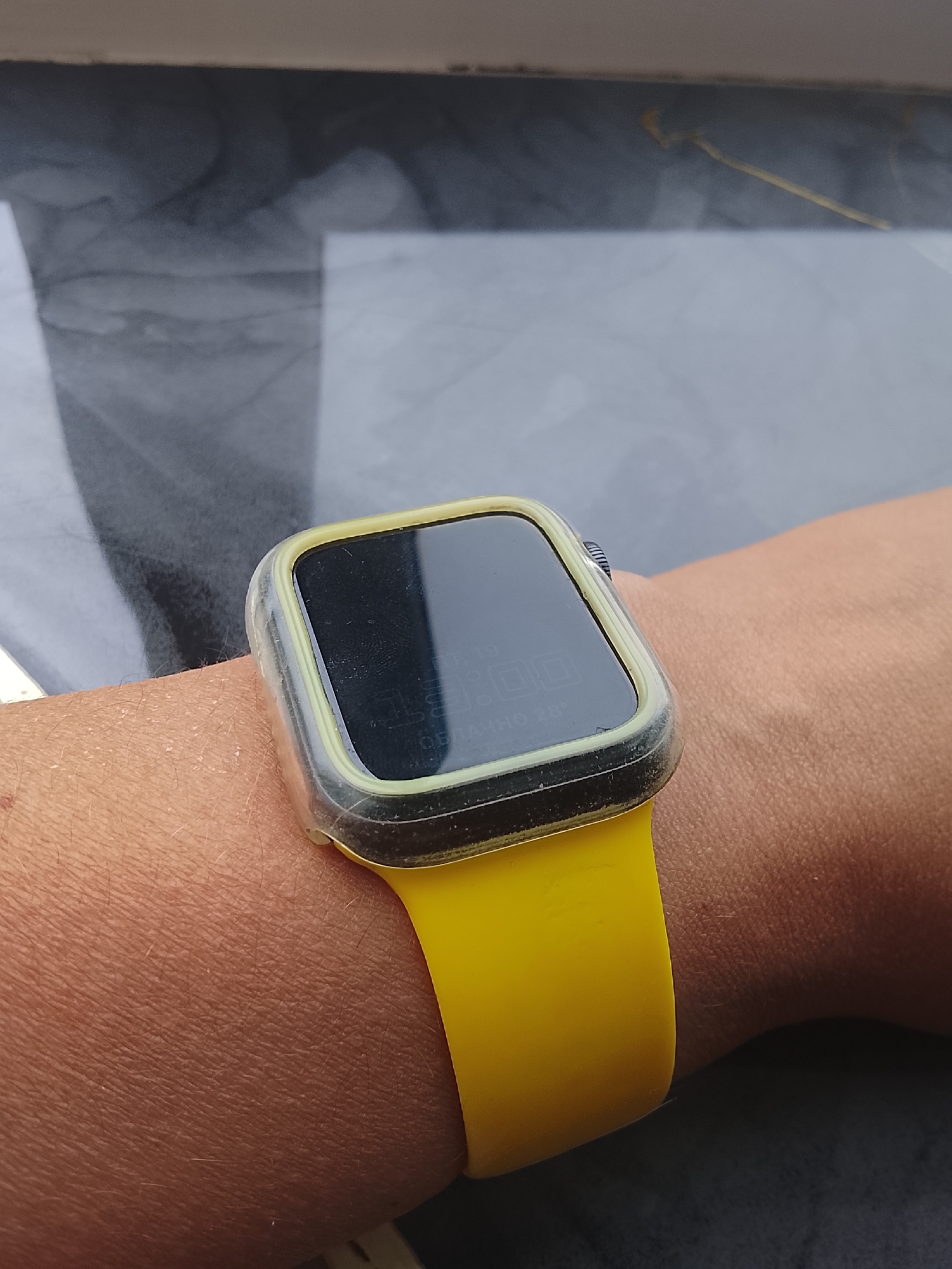 A close-up shot captures a person's wrist wearing a yellow smartwatch with a transparent protective case. The smartwatch displays a digital time of "19:00" and what appears to be "44H0 28°". The background is out of focus, showing a dark, possibly textured surface. The image was taken in Almaty, Kazakhstan.