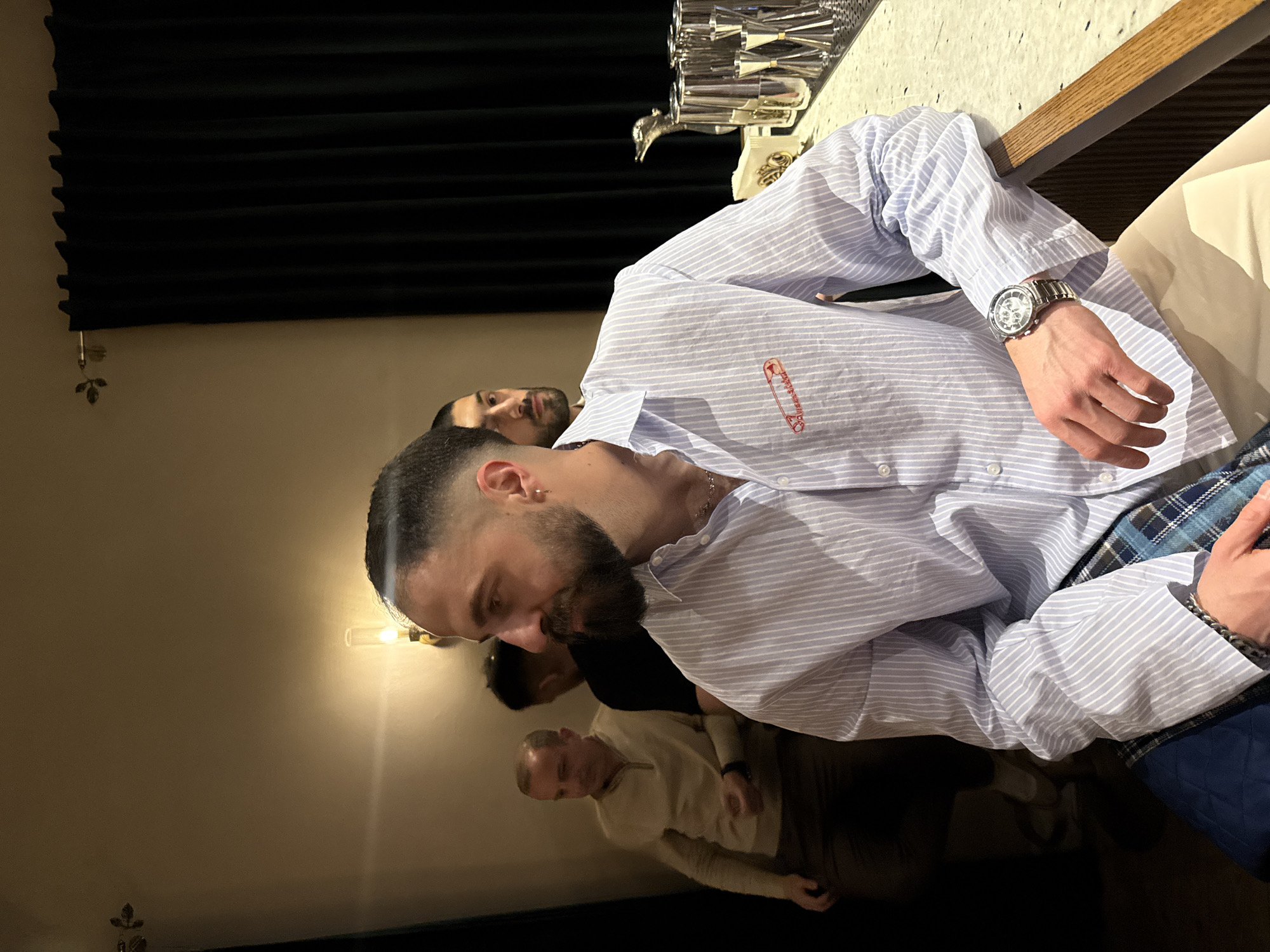 The image depicts a group of people in an indoor setting, likely a restaurant or bar. The primary subject is a man with a beard and short hair, wearing a light blue striped shirt with a safety pin detail. He is seated at a table, and his watch is visible on his left wrist. Behind him, another man is visible, partially obscured. In the background, there is a wall with a light fixture and dark drapery. On the table, there are glassware and a white countertop. A third man is partly visible on the lower part of the image, wearing a white top and with short, light-colored hair.
Given the setting and the mention of Yerevan, Armenia, it's reasonable to infer this is a social gathering in a restaurant or bar in that city.