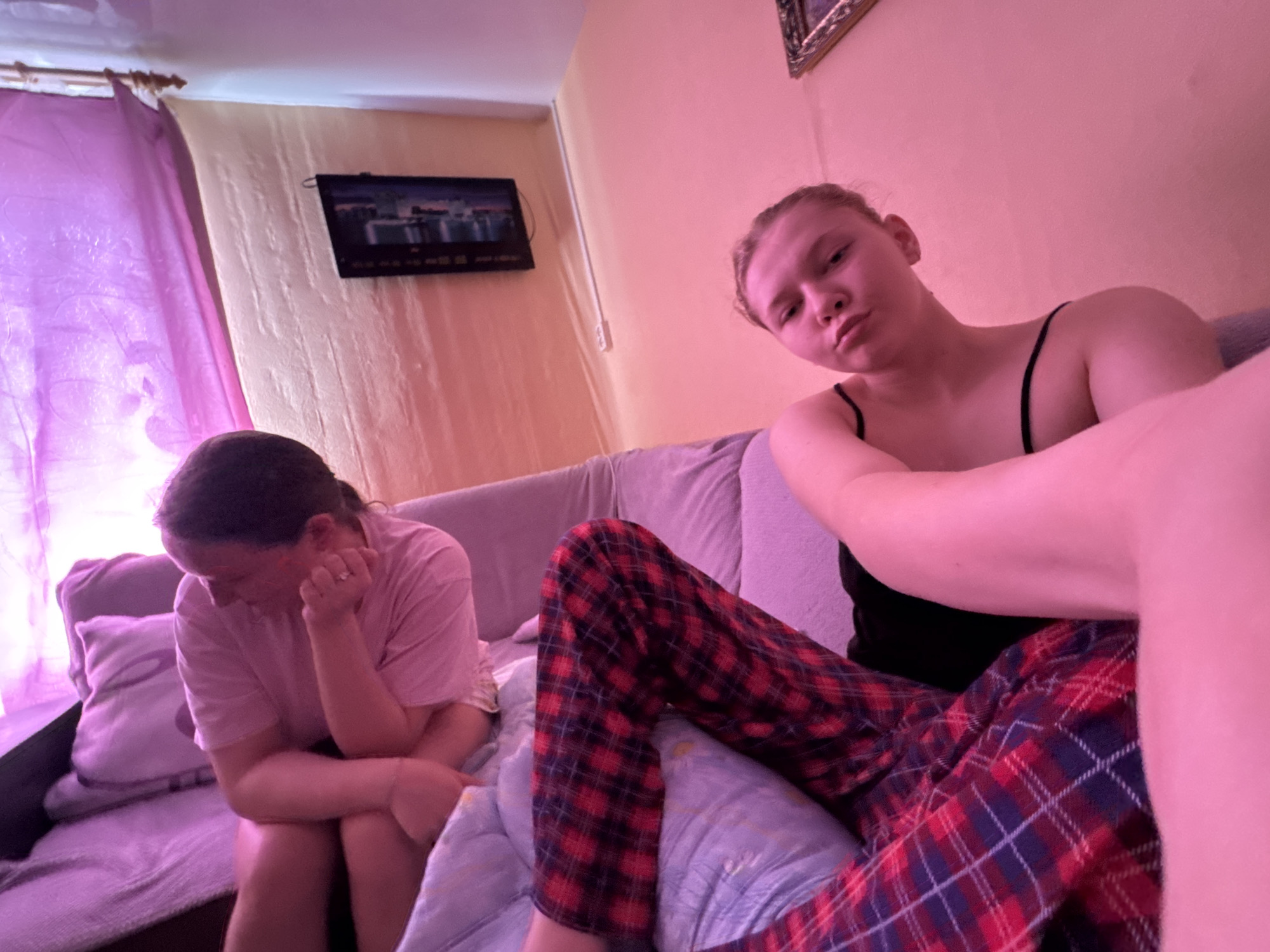 Two individuals are present in a room. One person sits on a couch, wearing a pink t-shirt and appearing pensive with their head bowed and hands near their face. The second person, positioned closer to the camera, is wearing a black tank top and plaid pants, sitting with their legs extended. A television is mounted on the wall. The setting is assumed to be indoors, on a couch, in what appears to be a domestic environment in Бородулино, Russia, given the instruction to include the location. The image does not provide information to confirm that the image was taken in this location. There is no information to prove that it was taken in the mentioned city. The presence and location cannot be confirmed.