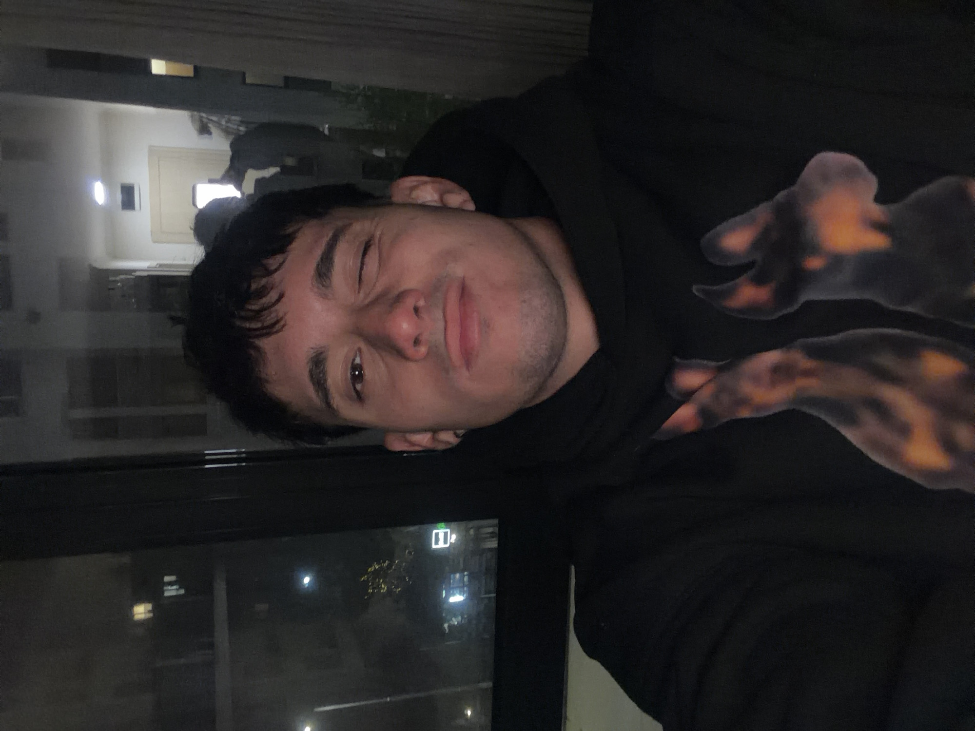 A man is looking at the camera, smiling with one eye closed, wearing a black hoodie with a blurry, dog-like print on the chest. He appears to be inside, near a window. Outside the window, lights and buildings are visible, suggesting a nighttime urban environment. There is an illuminated emergency exit sign visible on the right of the window. I am unable to determine the location in Sofia, Bulgaria from the image.