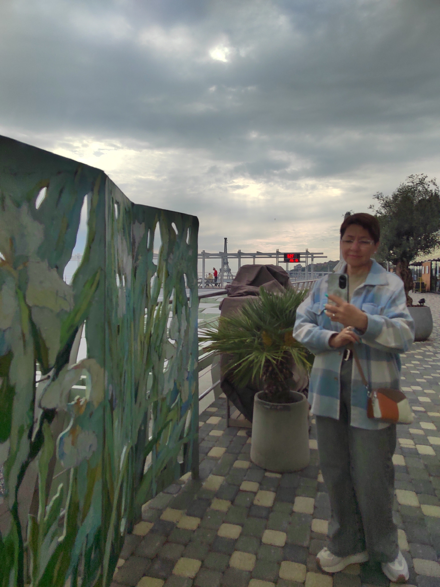 A woman stands on a paved walkway, holding a smartphone as if taking a selfie or photo. She wears eyeglasses, a patterned button-down shirt, pants, and sneakers, with a crossbody bag. Behind her, a potted plant and a cutout metal artwork depicting floral designs are visible. In the background, a structure reminiscent of the Eiffel Tower, a digital display showing '02:29', and a cloudy sky form the backdrop. The location appears to be a waterfront area in the city of Unknown, Unknown.