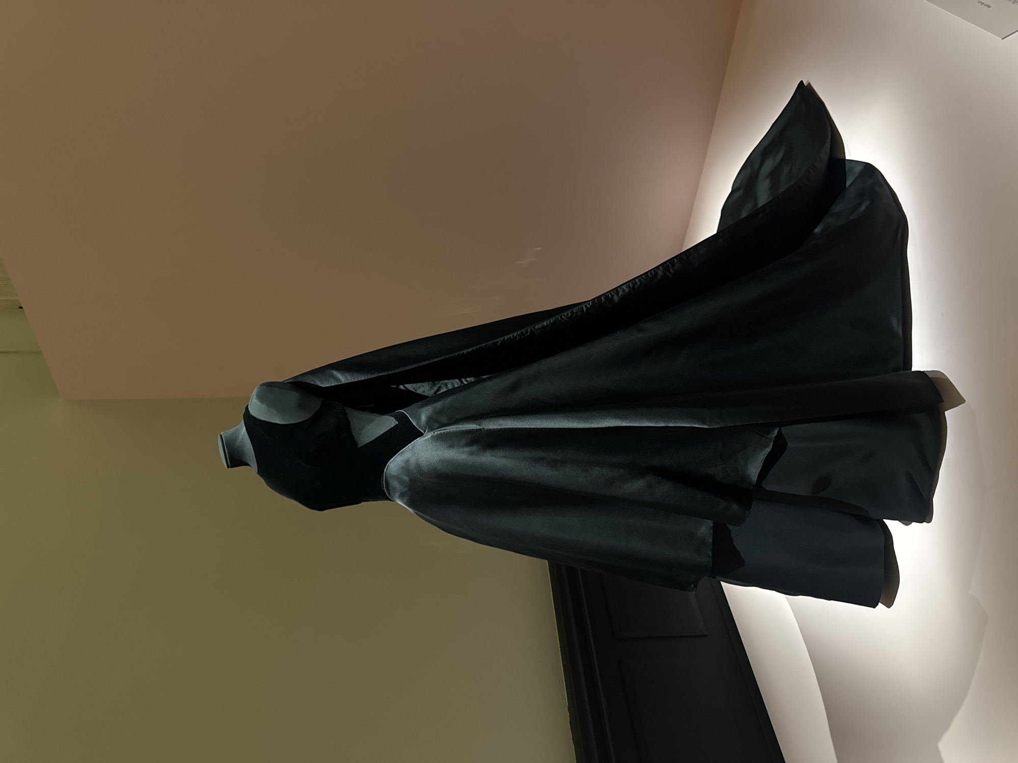 A black garment, possibly a robe or cloak, is displayed on a mannequin or stand within an interior space. The lighting is directed from the right side, casting a strong shadow on the wall behind the garment. The walls are painted in varying shades of beige. The image was taken inside a museum in Paris, France.
