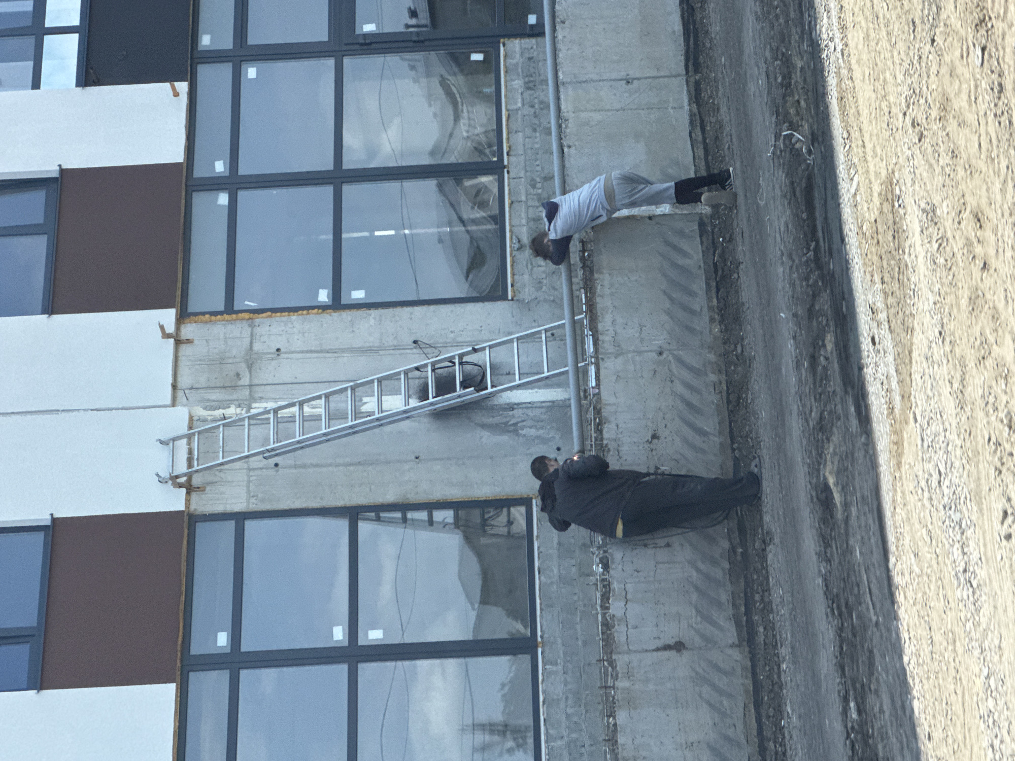 The image shows a construction site in Mikhaylovsk, Russia. Two individuals are present, standing near a concrete wall. The person on the left is wearing a dark jacket and dark pants, facing away from the camera and looking down. The person on the right is wearing a light-colored hooded sweatshirt and light-colored pants, leaning over a metal railing with their head down. A silver ladder is propped against the wall behind them, extending up towards a row of large windows. The environment appears to be a partially constructed building with rough concrete surfaces and a dirt and gravel ground.