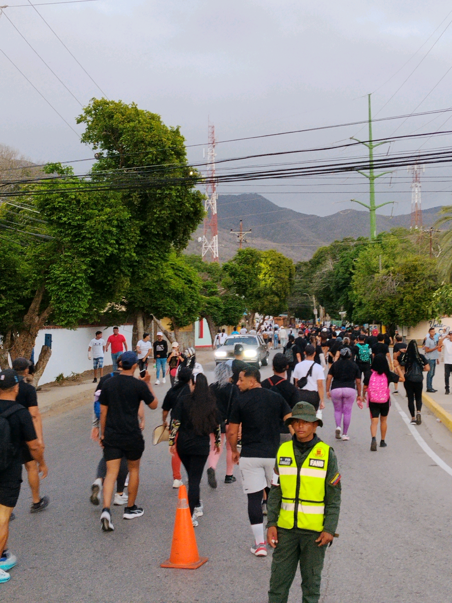 The image depicts a street scene in El Valle del Espíritu Santo, Venezuela, likely during an organized event or gathering. A large group of people are walking along a paved road, many dressed in dark clothing, suggesting a unified participation. Several individuals are visible in the foreground and mid-ground, including a security officer in a bright yellow vest and dark uniform standing at the right. The officer appears alert and is facing forward. The road is flanked by trees and buildings, with utility poles and power lines overhead. In the background, mountains are visible, and several communication towers pierce the sky. The presence of a traffic cone indicates some form of road management or event demarcation. The overall atmosphere appears to be one of a public assembly under a moderately overcast sky.