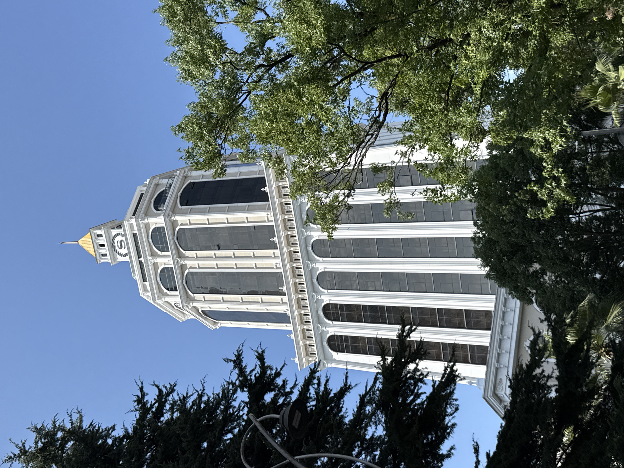 This is a low-angle, full-frame shot of the Batumi Alphabet Tower, a skyscraper located in Batumi, Georgia, on a clear, sunny day. The tower is white with a gold-colored cap and a clock face near the top. It is partially obscured by lush green trees in the foreground and on the right side of the frame. A dark-colored street lamp is visible on the left, also partially obscured by trees. The sky is a bright, cloudless blue.