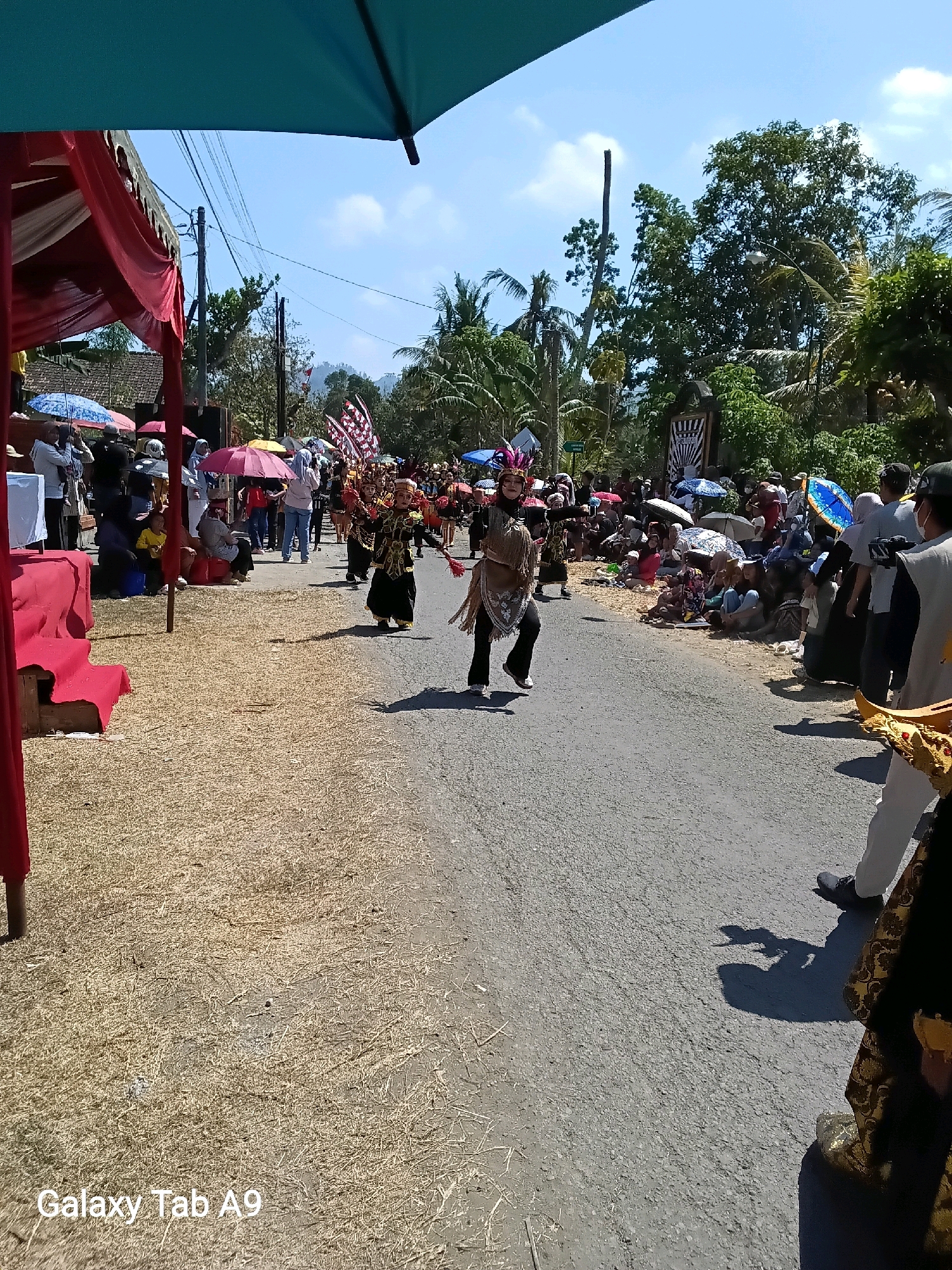 In Ngadirenggo, Indonesia, a street parade is underway under a bright, sunny sky. The road is lined with spectators, many holding umbrellas for shade, who are gathered on both sides of the asphalt. On the left, a red and white tent structure is set up, with some people seated and others standing around it. In the center of the road, two performers in traditional costumes are dancing, one in a black and gold outfit with a headdress and the other in a beige costume with fringe details. The ground is covered with dry grass and straw. Trees and buildings are visible in the background, with power lines strung across the sky. A person holding a phone or camera is in the foreground on the right, and another person in elaborate attire is partially visible on the far right. The scene suggests a cultural event or celebration taking place in a rural or semi-rural setting.