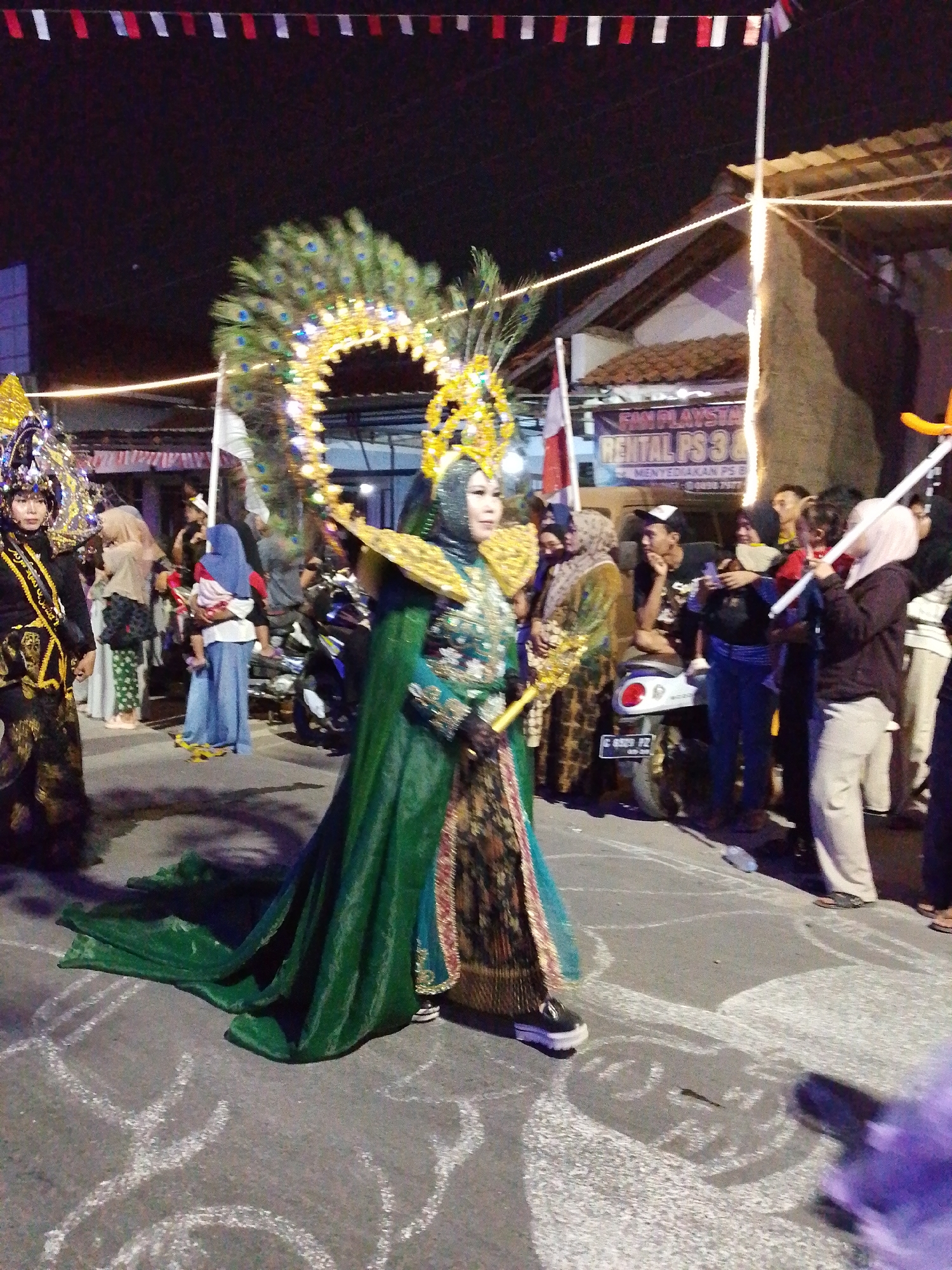 In the city of Pagongan, Indonesia, a nighttime street parade is captured in this image. The focus is on two individuals in elaborate costumes. The person in the foreground is adorned in a vibrant green cape with intricate gold embellishments, a jeweled hijab, and a magnificent peacock feather headdress. To their left, another participant wears a dark, sequined outfit with a similarly ornate headdress and a golden sash. Both are walking down a street illuminated by overhead lights and string lights. In the background, onlookers are gathered, some on motorcycles, and buildings with awnings line the street. A banner advertising "RENTAL PS 3 & 4" is visible on one of the buildings. The street itself is marked with white chalk drawings, suggesting a festive or artistic element to the event. The overall atmosphere is one of celebration and cultural display.
