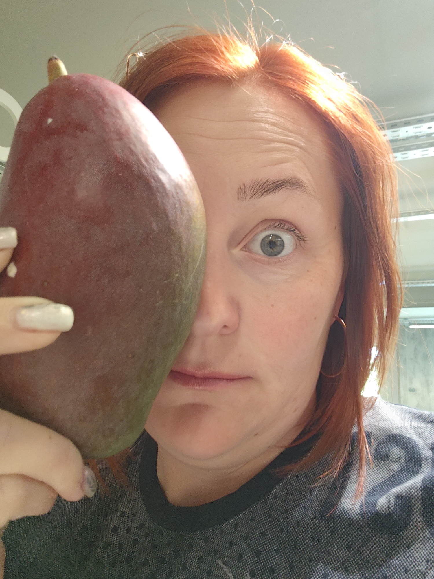 A reddish-haired woman with light skin holds a large mango in front of her face, obscuring half of her features. The woman's left eye is visible, and she is wearing a dark-colored patterned shirt. Her fingernails are light in color. The background appears to be an interior space with metal shelving. The location of Марковский, Russia, is not discernible from the image.