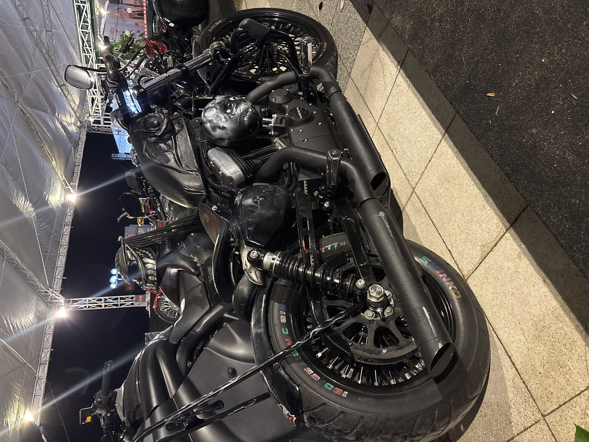 The image shows a close-up of a custom black motorcycle parked on a tiled surface, adjacent to a carpeted area. The bike has a large front tire with the brand "SHINKO" visible in multicolored lettering. The motorcycle features blacked-out components, including the engine, exhaust pipes, and spoked wheels. In the background, there is a structure covered with white material and scaffolding visible, potentially part of an event setup. The location could be in a city setting. Based on the prompt, this is in Nha Trang Ward, Vietnam. The scene is likely taken at night or in low light, considering the illumination from the structure in the background.