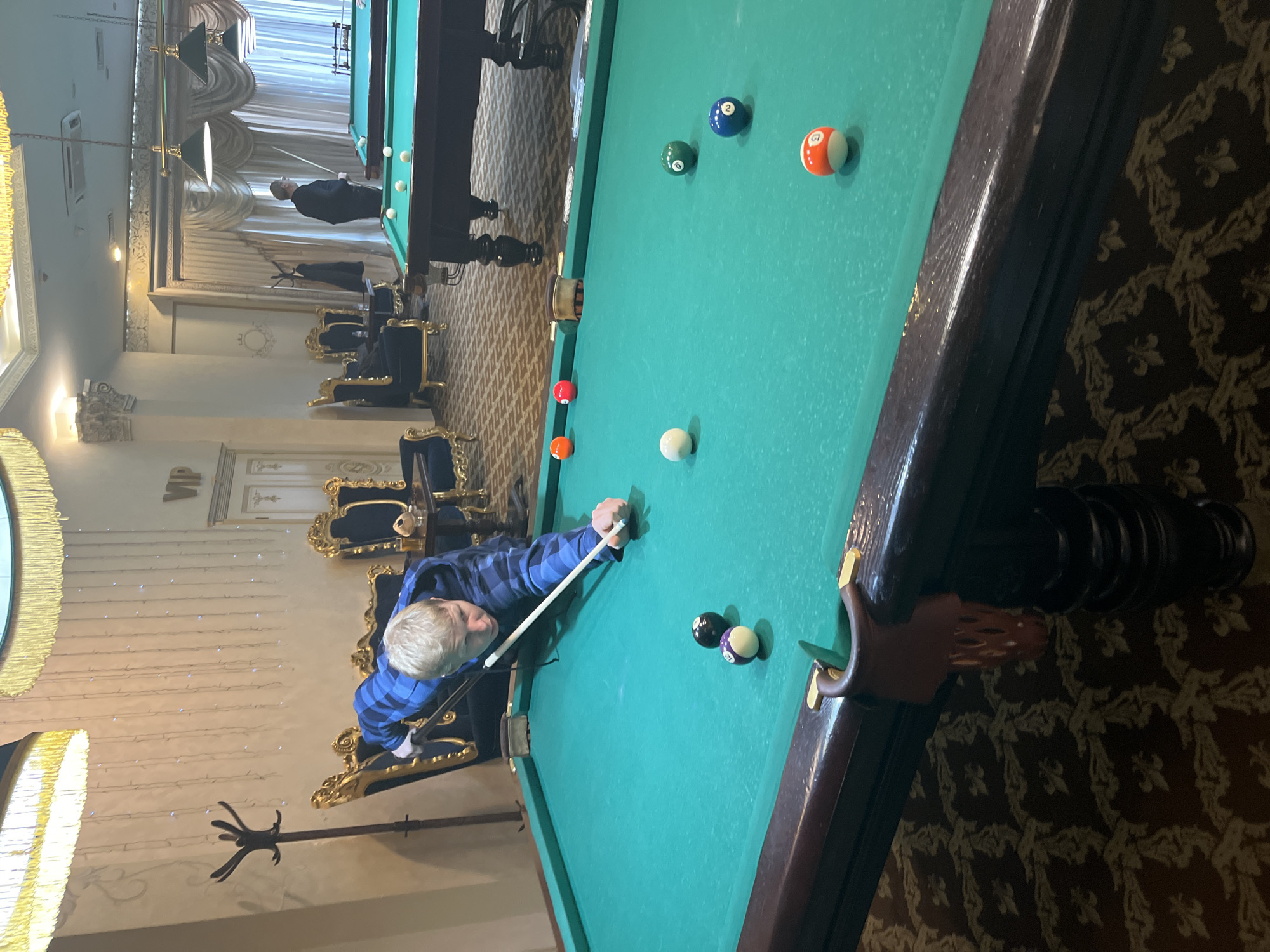 The image shows a man playing billiards in an ornately decorated room. The man, wearing a blue plaid shirt, is positioned at the end of a billiard table, cue in hand, aiming at the balls. Several billiard balls are scattered across the green surface of the table. In the background, there are other billiard tables with people playing, along with ornate chairs and light fixtures. The room's decor suggests a luxurious setting. The location is Sochi, Russia.