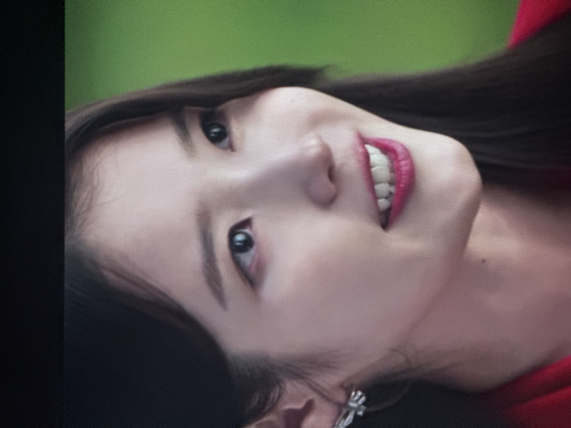 The image captures a close-up portrait of a young East Asian woman, rotated to appear on her side. She is smiling broadly, with her lips painted in a vibrant fuchsia or red hue, revealing her upper teeth. Her dark hair frames her face. She wears a silver-colored, possibly diamond-encrusted, earring on her visible earlobe and a red garment, likely a top or dress. Her expression is one of happiness or amusement. The background is softly blurred, displaying a bright green color on the upper and right sides, suggesting outdoor foliage or a park-like setting during daytime. A dark, vertical element occupies the left edge of the frame. The lighting on her face is soft and even. No text is visible in the image. The setting is Budva, Montenegro.