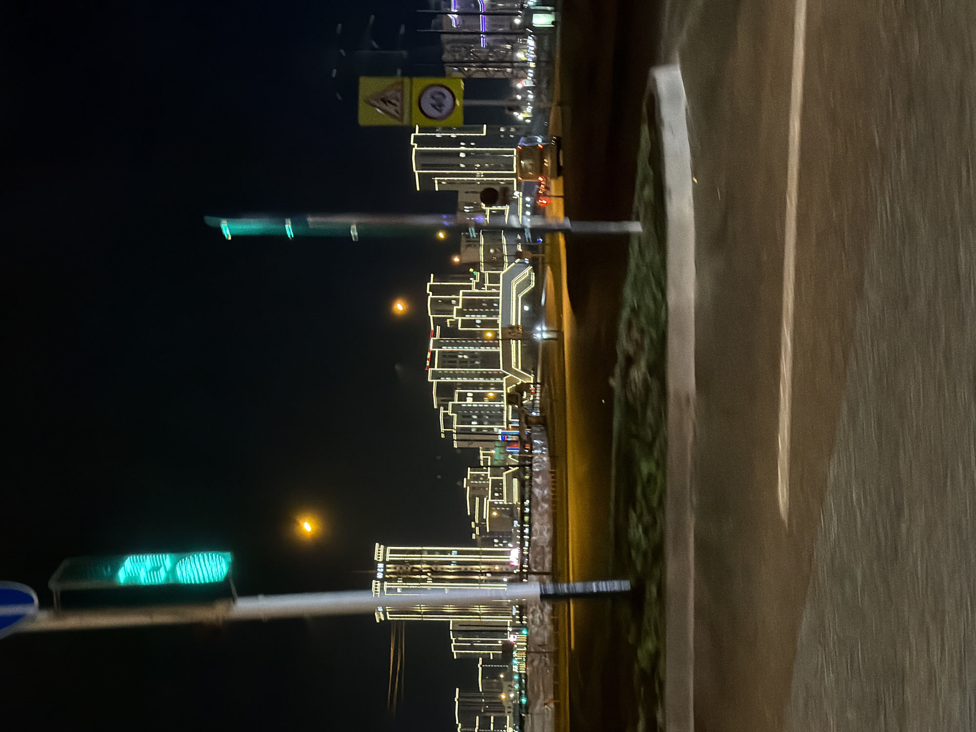 The image depicts a night scene in an urban environment. The foreground features a road or pavement, slightly blurred, with a concrete barrier and a strip of greenery. Further in the distance, there are several buildings illuminated with decorative lights, outlining their architectural features. A yellow road sign is also visible. In the lower portion of the frame, a traffic light with a green signal is noticeable. The overall lighting is dim with a dark sky in the background.
