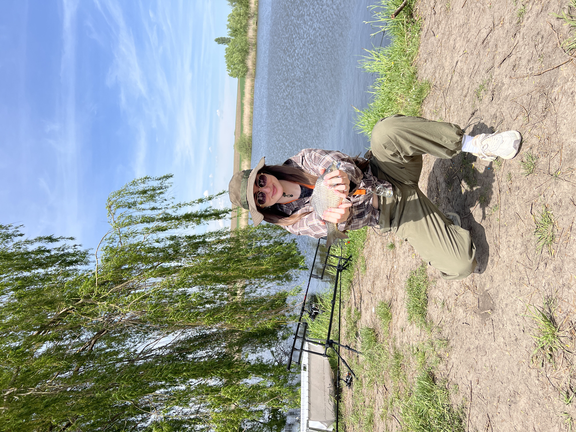 A woman in a hat and sunglasses is kneeling near a body of water, holding a fish. She is wearing a plaid shirt and olive-green pants and white sneakers. There is a fishing rod stand nearby. The location appears to be Romanove Selo, Ukraine, based on the presence of water and vegetation. The weather seems sunny. The focus is on the woman and the fish she is holding.