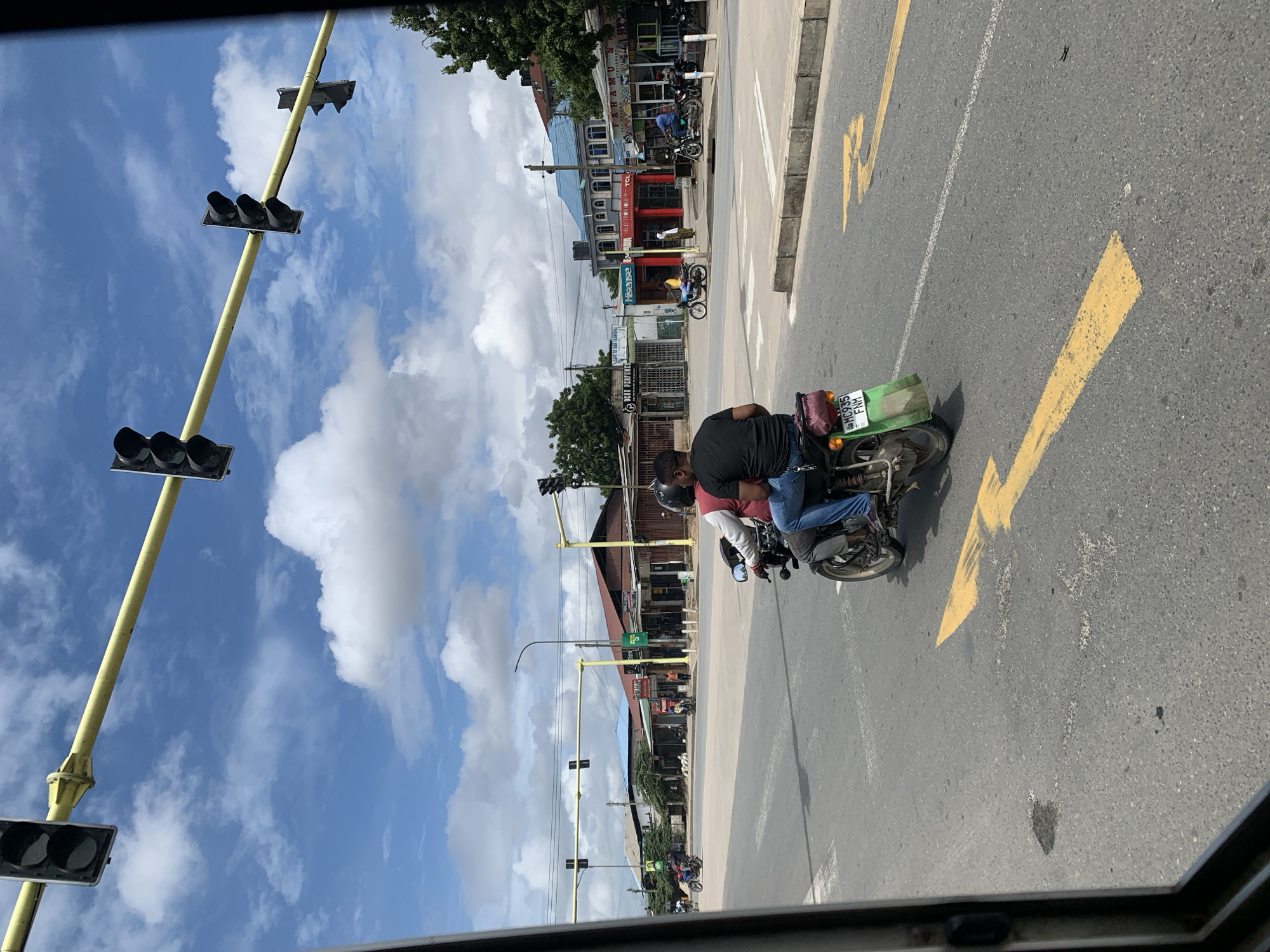 A photo taken from inside a vehicle shows a street scene in Dar es-Salaam, Tanzania, on a sunny day with a few clouds.
A motorcycle with two riders is in the foreground on the right side of the frame. The rider in the back is wearing a black shirt, and the driver is wearing a red and white shirt. Both are wearing jeans and helmets. The motorcycle has a green carrier on the back and a Tanzanian license plate.
The street has yellow painted directional arrows. There are several traffic lights along the street. Buildings line the street on both sides, with a mix of shops and commercial establishments. The color blue is prominent in the building facades. Several other people and motorcycles are visible further down the street.