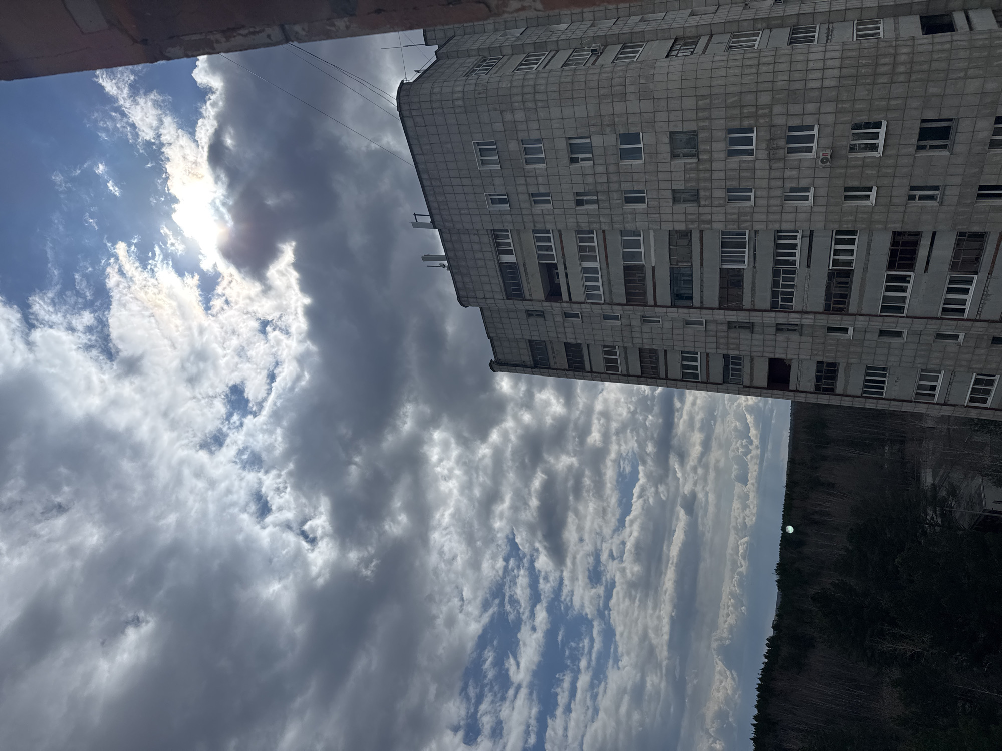 This image captures a stark urban and natural landscape in Chaikovsky, Russia, viewed from a high vantage point, possibly a window. The scene is dominated by a tall, grey, multi-story building, a dramatic sky, and a distant forest.
The main subject is a large, concrete apartment building occupying the right side of the frame. It exhibits a blocky, grid-like facade with numerous rectangular windows, many featuring white frames, others darker. An external unit, possibly an air conditioner, is visible on one of the upper floors. The building has a subtly curved corner near its top, suggesting a specific architectural style common in post-Soviet environments.
The sky fills the left and central portions of the image, showcasing a dynamic interplay of light and shadow. Bright sunlight bursts through a bank of luminous white clouds, creating a strong glare and highlighting their edges. Adjacent to these are heavier, darker grey clouds, indicating a partly cloudy or clearing weather condition. Patches of lighter blue sky are visible in some areas.
In the lower distance, a dense, dark green and brown forest stretches horizontally across the horizon line. Just above the treeline, a faint, circular, light-colored object is discernible, its identity unclear but possibly a distant structure or an optical artifact. Thin utility wires are visible against the sky near the top edge of the building. The very top of the image is framed by a dark, reddish-brown structure, possibly an overhang or part of another building.
The strong, bright sun breaking through the clouds suggests it is daytime, possibly morning or late afternoon, creating a dramatic and somewhat somber atmosphere. No people or discernible text are visible in the image.