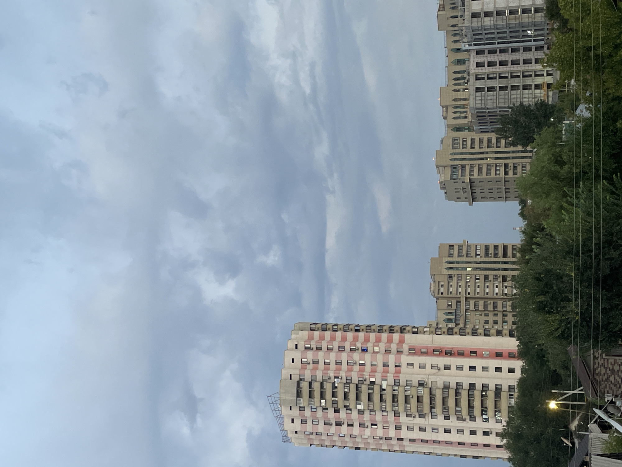 The image captures a street view in Almaty, Kazakhstan, featuring several high-rise apartment buildings against a backdrop of a cloudy sky. In the foreground, lush green trees partially obscure smaller residential structures and a streetlamp. Dominating the scene are three prominent apartment buildings of varying architectural styles and stages of completion.
The leftmost building is a tall, slender structure with a distinctive pink and white striped facade, punctuated by numerous windows and balconies. To its right stands another mid-rise building, characterized by its beige coloration and ornate, arched window details. Adjacent to this, a multi-story building appears to be under construction, its concrete frame and exposed windows revealing its unfinished state. The overall atmosphere is subdued, likely due to the overcast sky. No individuals or specific activities are discernible in the image.