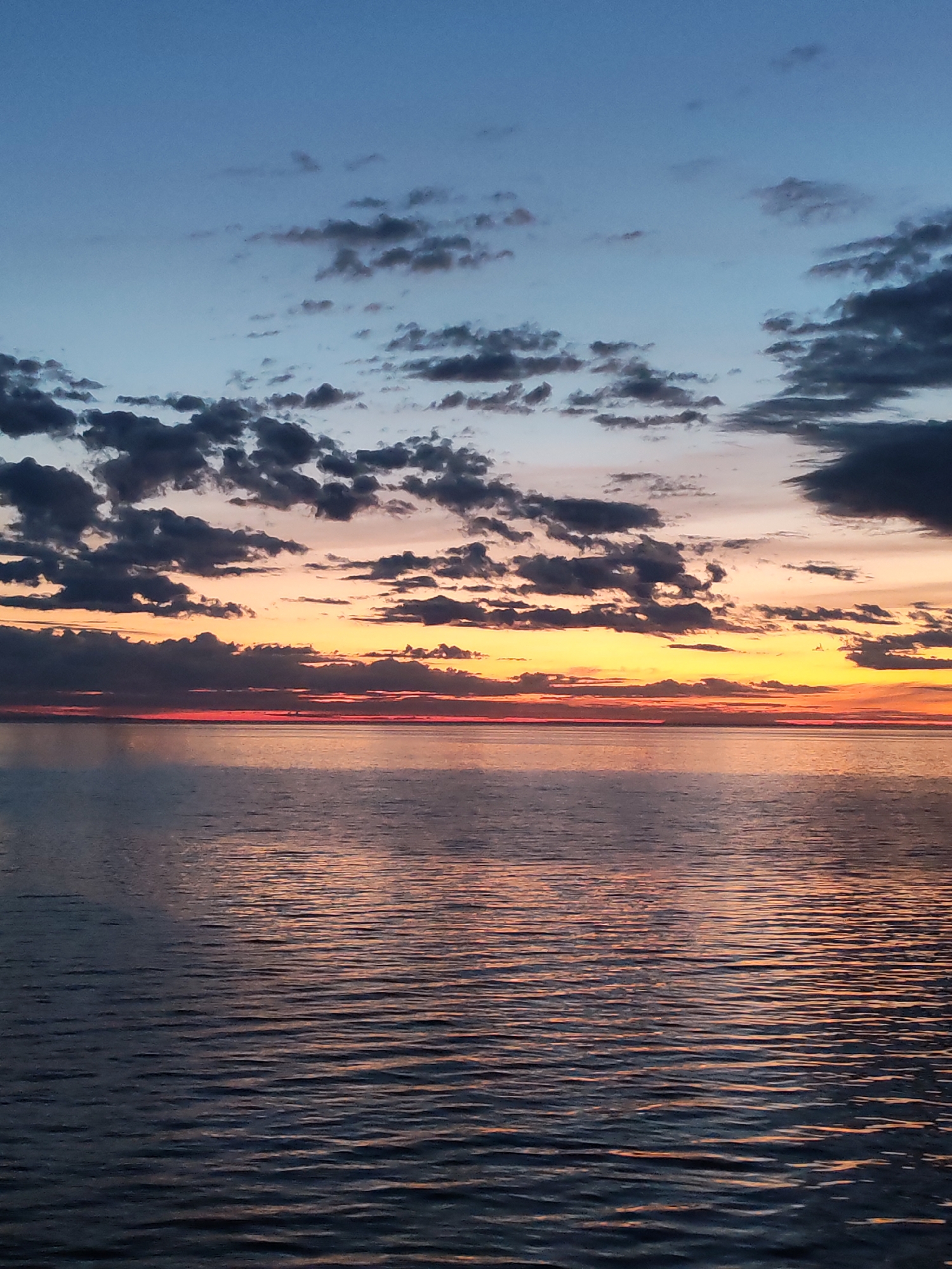 The image depicts a sunset over a body of water. The sky is a gradient of colors, transitioning from blue at the top to orange and yellow near the horizon. There are dark, fragmented clouds scattered across the sky, particularly near the horizon. The water reflects the colors of the sky, creating a shimmering effect with ripples on the surface. The location is Unknown, Unknown. No people, specific subjects, objects, actions, or interactions are discernible beyond the natural environment of the sunset and water.
