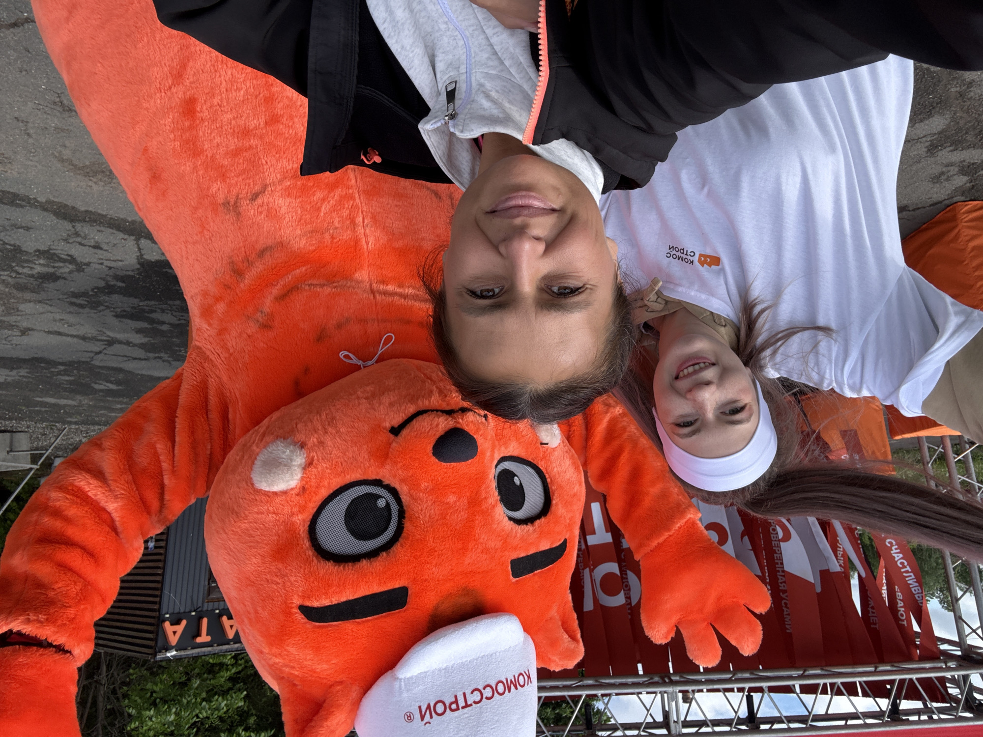 Two women are posing for a selfie with a large orange mascot of a cat wearing a white hat labeled "KOMOCCTPOЙ" at an event in Izhevsk, Russia. The woman on the left has dark hair, and is wearing a black jacket over a gray sweatshirt. The woman on the right has long brown hair, a white headband, and a white T-shirt. Behind them is a metal structure with red and white banners featuring text in Russian.