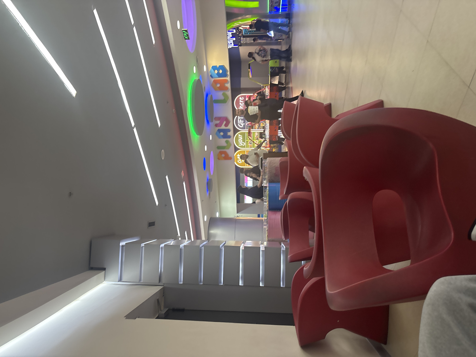 The image displays a brightly lit, modern indoor entertainment area, likely a play zone or arcade, situated in a commercial complex in Yerevan, Armenia. The scene is characterized by a sleek design and ample artificial lighting.
In the foreground and extending towards the middle-ground on the right, several rows of distinct red, molded plastic chairs are arranged, suggesting a waiting or seating area. The legs of a person are visible in the nearest chair. The ceiling is illuminated by numerous long, parallel LED light strips, providing even and intense white light.
The central feature of the background is a large, colorful, and illuminated sign that spells out "PLAY LAB" in varied, whimsical fonts and hues. Below this main sign, smaller illuminated signs read "your" and "GIFT here," along with additional words like "eat" and "play" which seem to be part of the establishment's branding or offerings. The ceiling in this section also features colorful, recessed circular and oval lights, contributing to a vibrant and playful atmosphere.
Several individuals, predominantly adults, are visible in the background, walking or standing near what appear to be arcade machines and interactive game stations. The floor is light-colored and tiled. On the left side of the image, there are modern, tiered white architectural elements or display shelves with integrated lighting, enhancing the contemporary aesthetic. The overall ambiance is one of leisure and activity within a well-maintained and engaging environment.