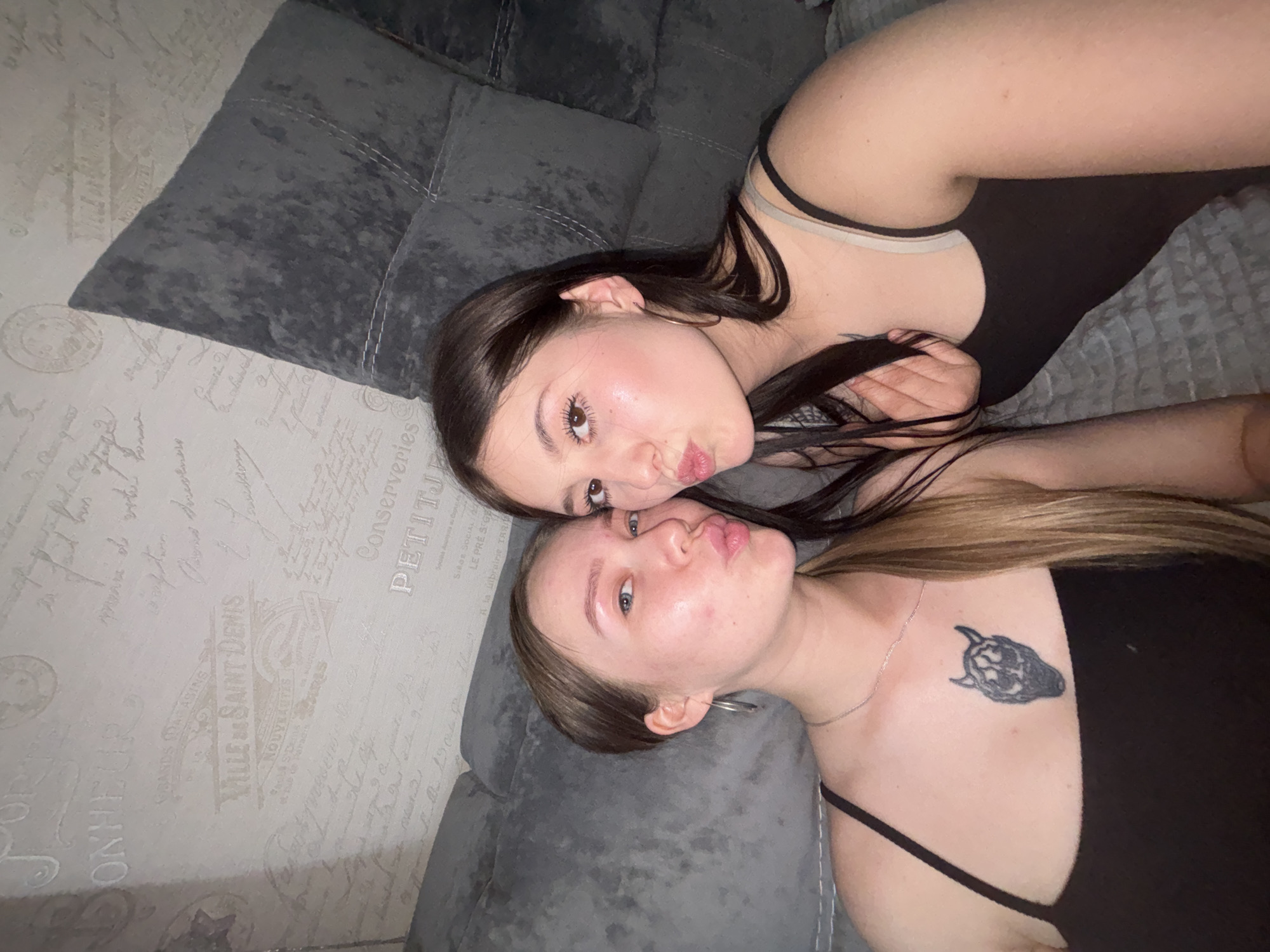 Two young women are lying next to each other on a grey couch, both wearing black tops. The woman on top has dark hair, while the other has blonde hair. Both are making a kissing face towards the camera. The background shows a wall with a patterned design. The image does not provide information related to Chaikovsky, Russia.