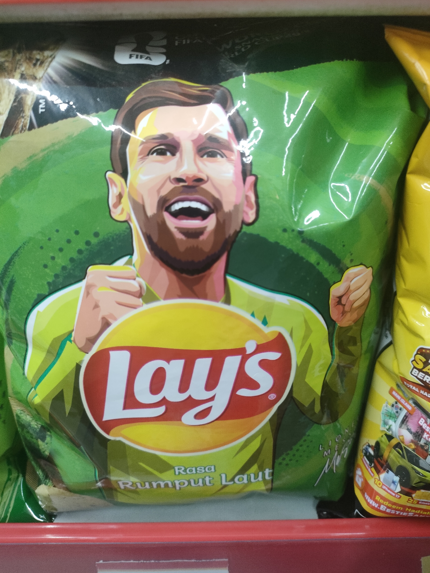 The image shows a close-up of a bag of Lay's potato chips featuring a cartoon illustration of Lionel Messi. The bag is predominantly green with Messi's face prominently displayed. The Lay's logo is in the center of the bag, with text below it in Indonesian. The bag appears to be on a shelf, with other products visible to the right. The lighting suggests an indoor setting, possibly a store. The background is a blur of green and yellow. There is no visible indication of the specific location being in Desa Banyumas, Indonesia, nor any discernible environmental details from that area.