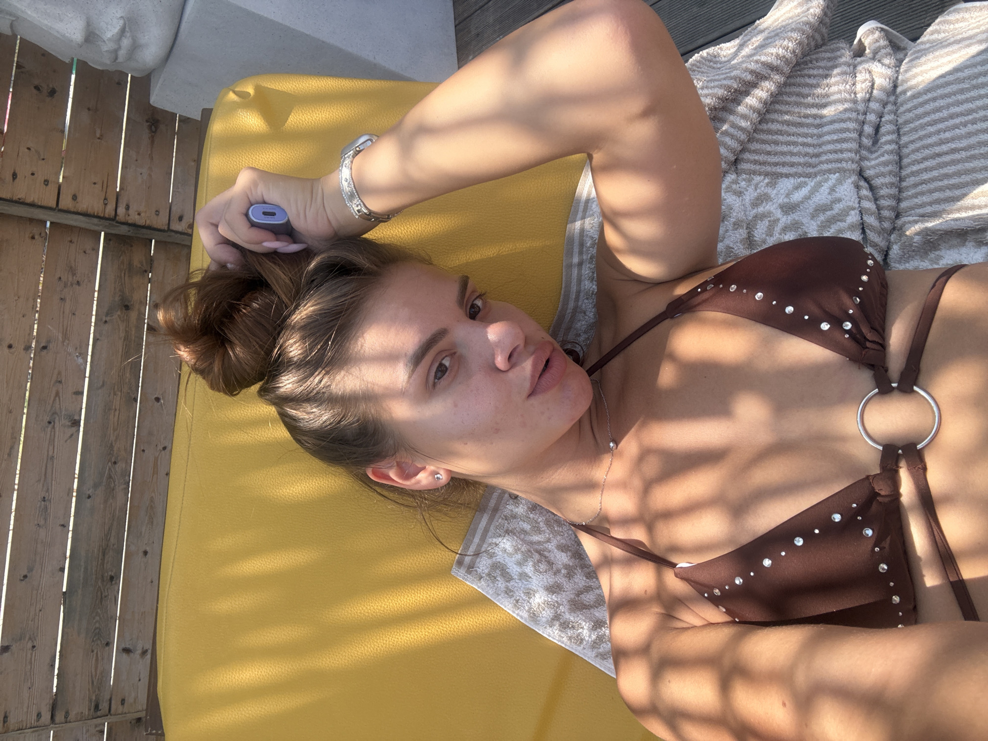 A young woman with her hair in a bun is lying on a yellow chaise lounge outdoors, possibly in a resort or beach setting, in Katerini Municipality, Greece. She is wearing a brown bikini with rhinestone embellishments and a silver ring detail. She is holding a small purple electronic device to her head with her right hand. A patterned towel is placed on the chaise lounge beneath her. Wooden slats form a fence or canopy above her, casting shadows on her and the lounge. A stone sculpture or bust is partially visible to the right of the frame. The scene is brightly lit by sunlight, suggesting a warm, outdoor environment.